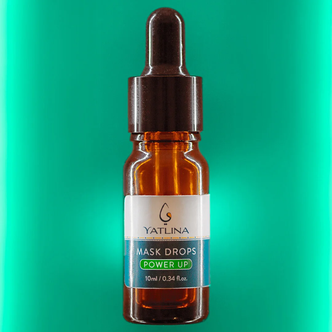 A small brown glass bottle with a black dropper cap labeled 'YATLINA MASK DROPS POWER UP 10ml / 0.34 fl. oz.' against a green background.