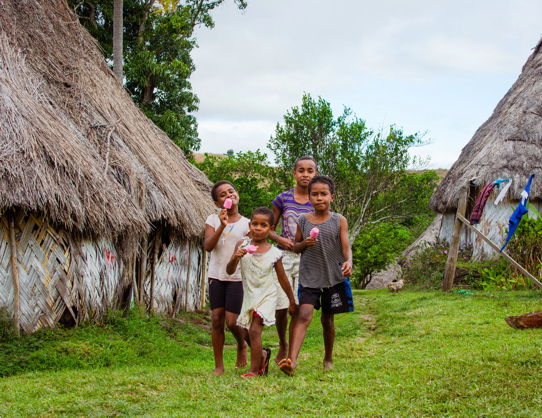 Four children walking on grass near rustic huts, holding pink ice pops, in a village setting with trees and cloudy sky.
