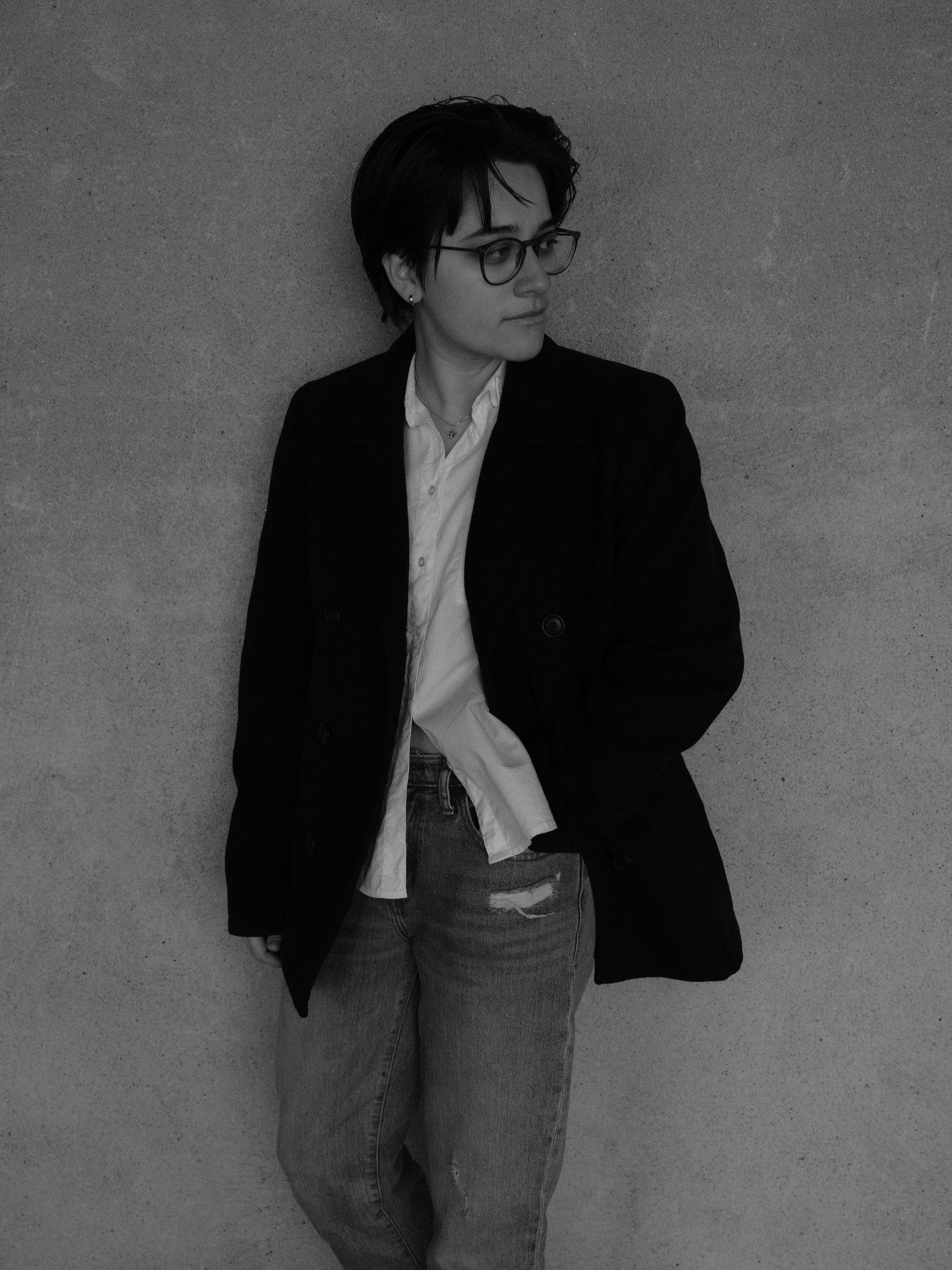 A woman with short dark hair, glasses, and earrings stands against a textured wall, wearing a white shirt, a dark blazer, and ripped jeans, looking to her right.