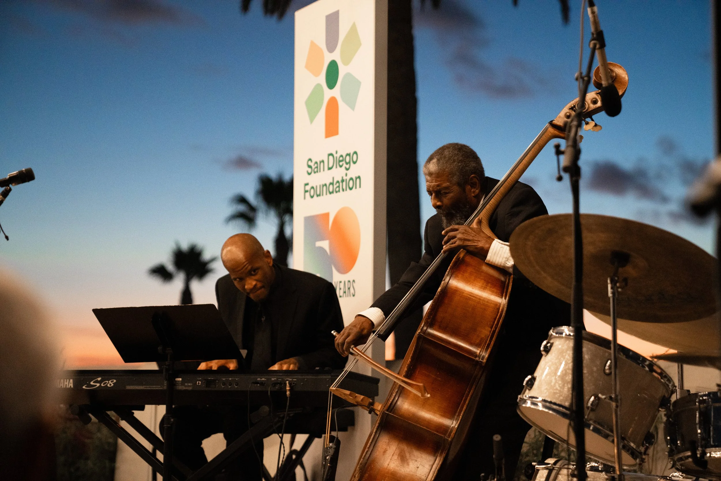 Two musicians perform outdoors at sunset, one playing a keyboard and the other playing a double bass, with a San Diego Foundation 50-year anniversary banner in the background.