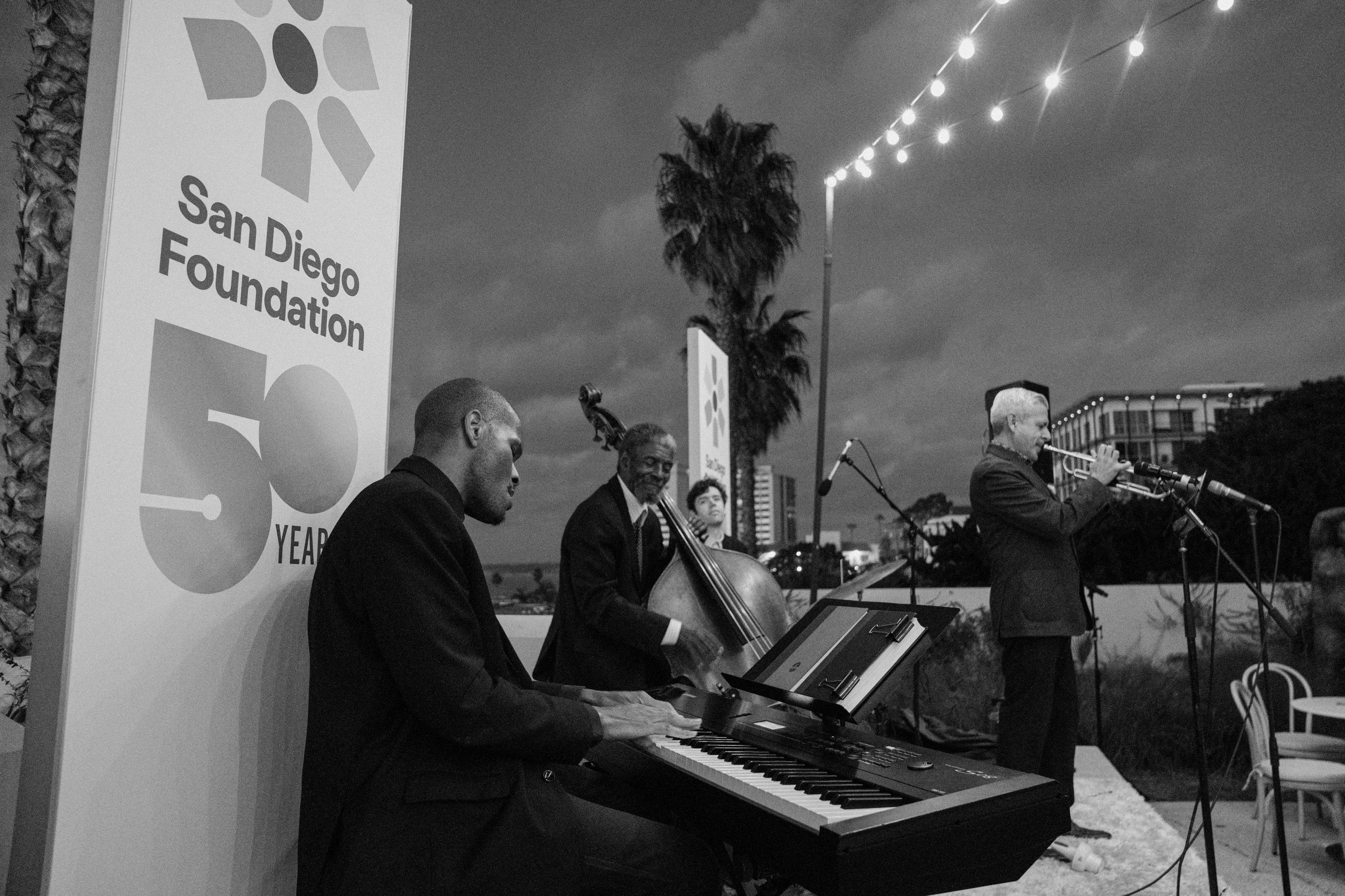 Musicians performing outdoors under string lights, with a sign reading "San Diego Foundation 50 Years" visible on the left.