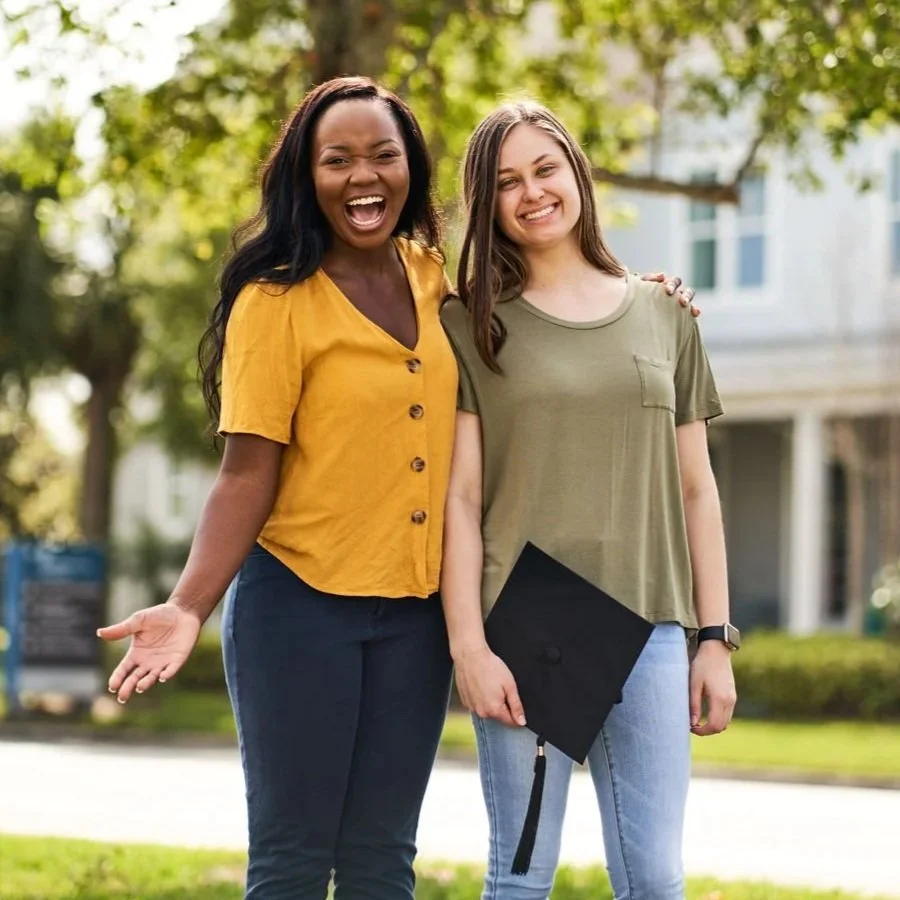 A coach standing next to a student outdoors with their arm around them. The student is holding a black graduation cap and they are both looking at the camera and smiling.