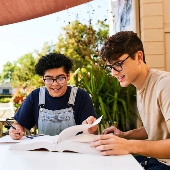 A coach and student sitting at a table outdoors. There is a book in front of them and the student is writing in a notebook.