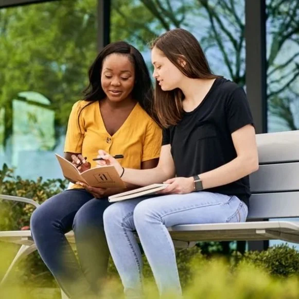 A coach and student sitting side-by-side on a bench outdoors, working together with notebooks, journals, and pens.
