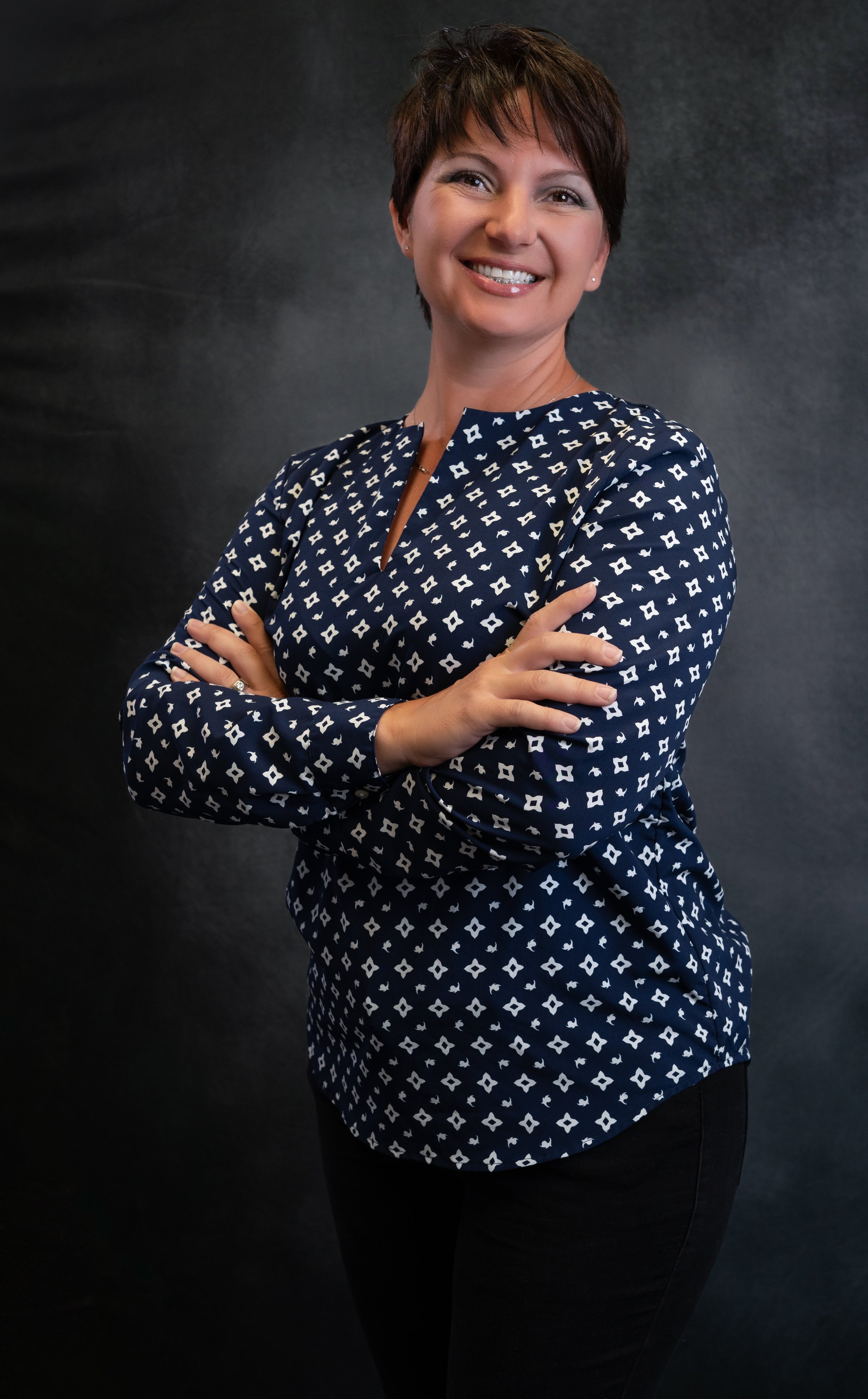 A smiling woman with short brown hair smiling with her arms crossed wearing a blue and white shirt