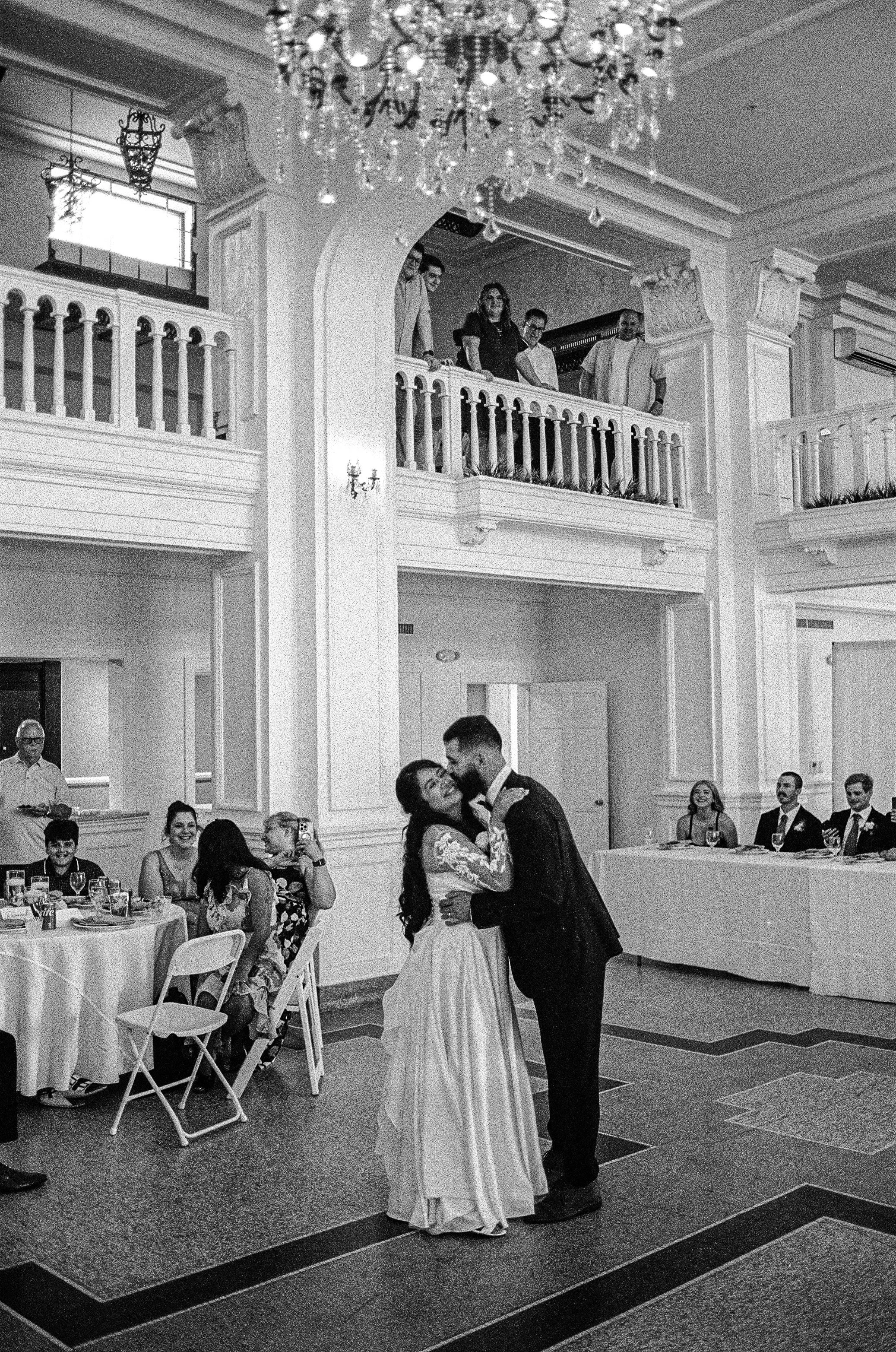 A wedding celebration with a couple dancing in the center of the room, surrounded by seated guests at decorated tables. The decor includes a chandelier, ornate columns, and a balcony with additional guests watching the couple dance.