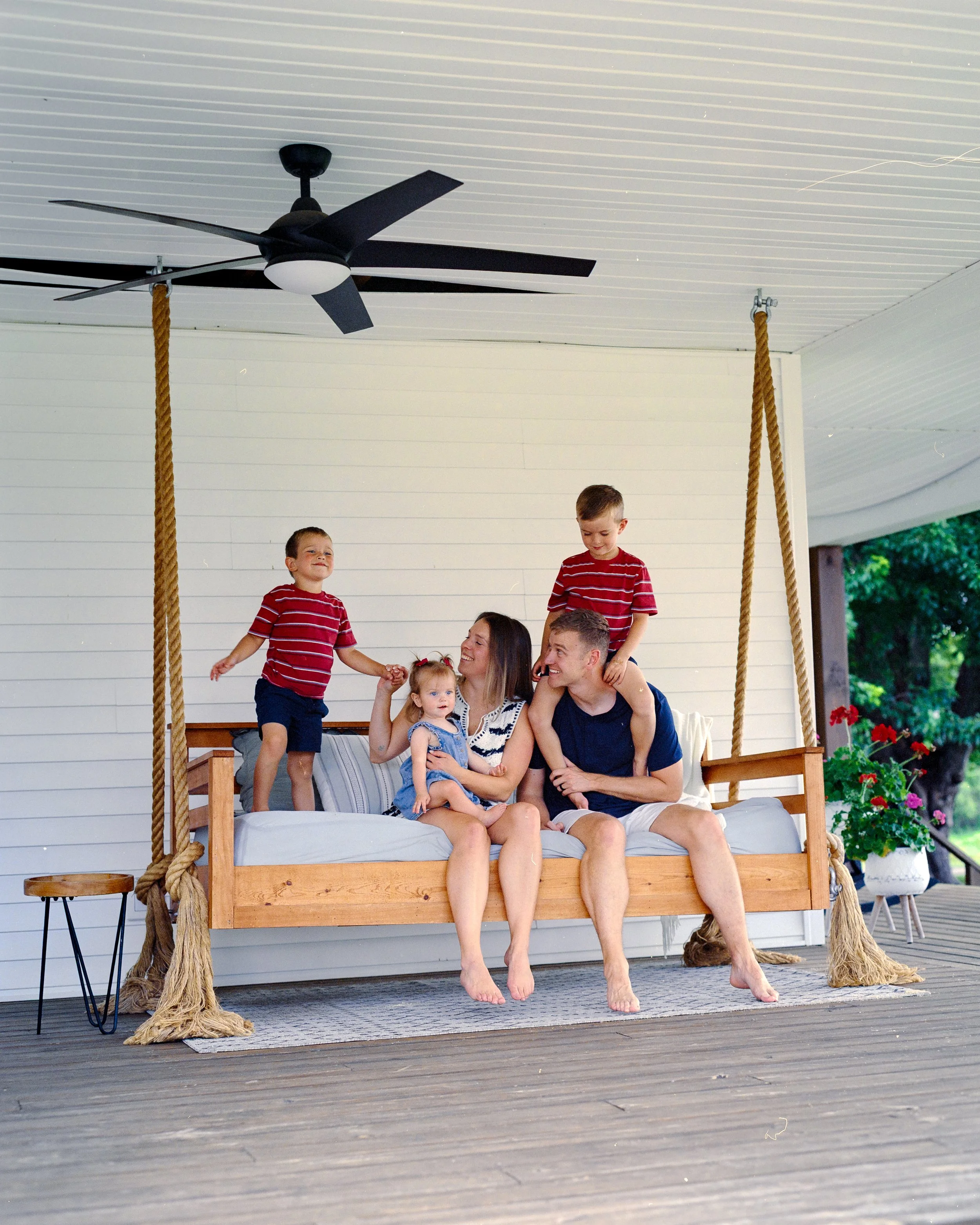 A family of six sitting on a bed that is in a porch swing setup, with four children and two adults. The children are all boys and are wearing red and white striped shirts; three are sitting and one is standing. The adults, a man and a woman, are sitting at the edge of the bed, holding a young girl with blonde hair. There are large ropes on either side of the swing, and a ceiling fan is visible above.