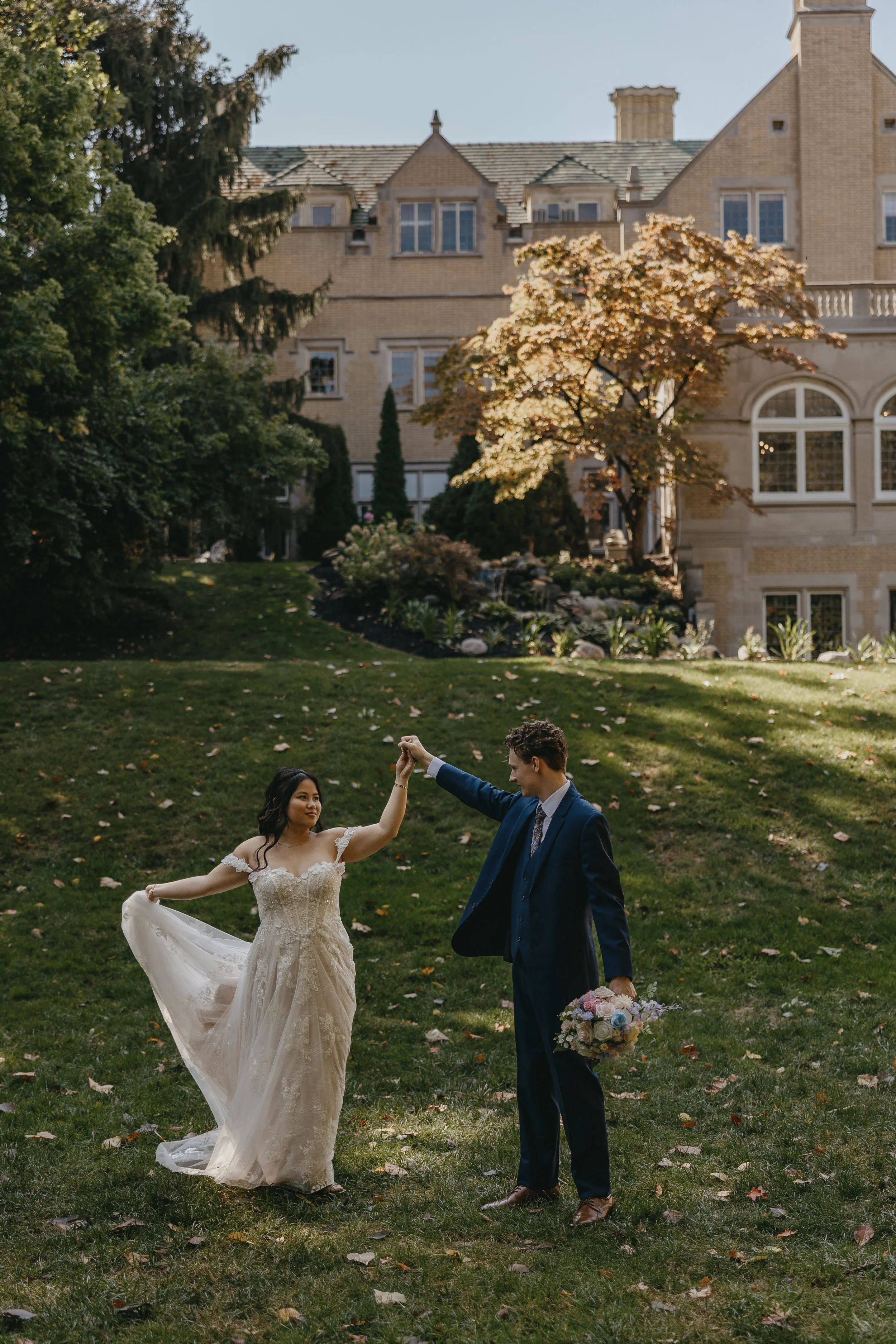 A bride and groom are dancing outdoors on a grassy lawn with a large historic house and trees in the background. The groom is holding a bouquet of flowers and the bride is wearing a lace wedding dress, while the groom is dressed in a dark suit.