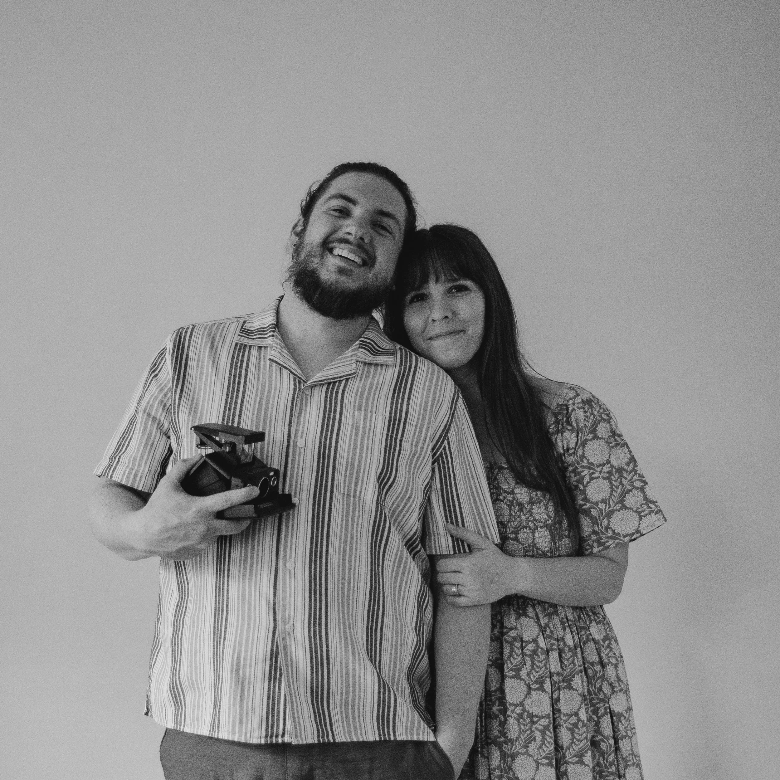 Two smiling people, one man with a beard and long hair, wearing a striped shirt, and one woman with long hair and bangs, wearing a patterned dress, standing closely together against a plain background.