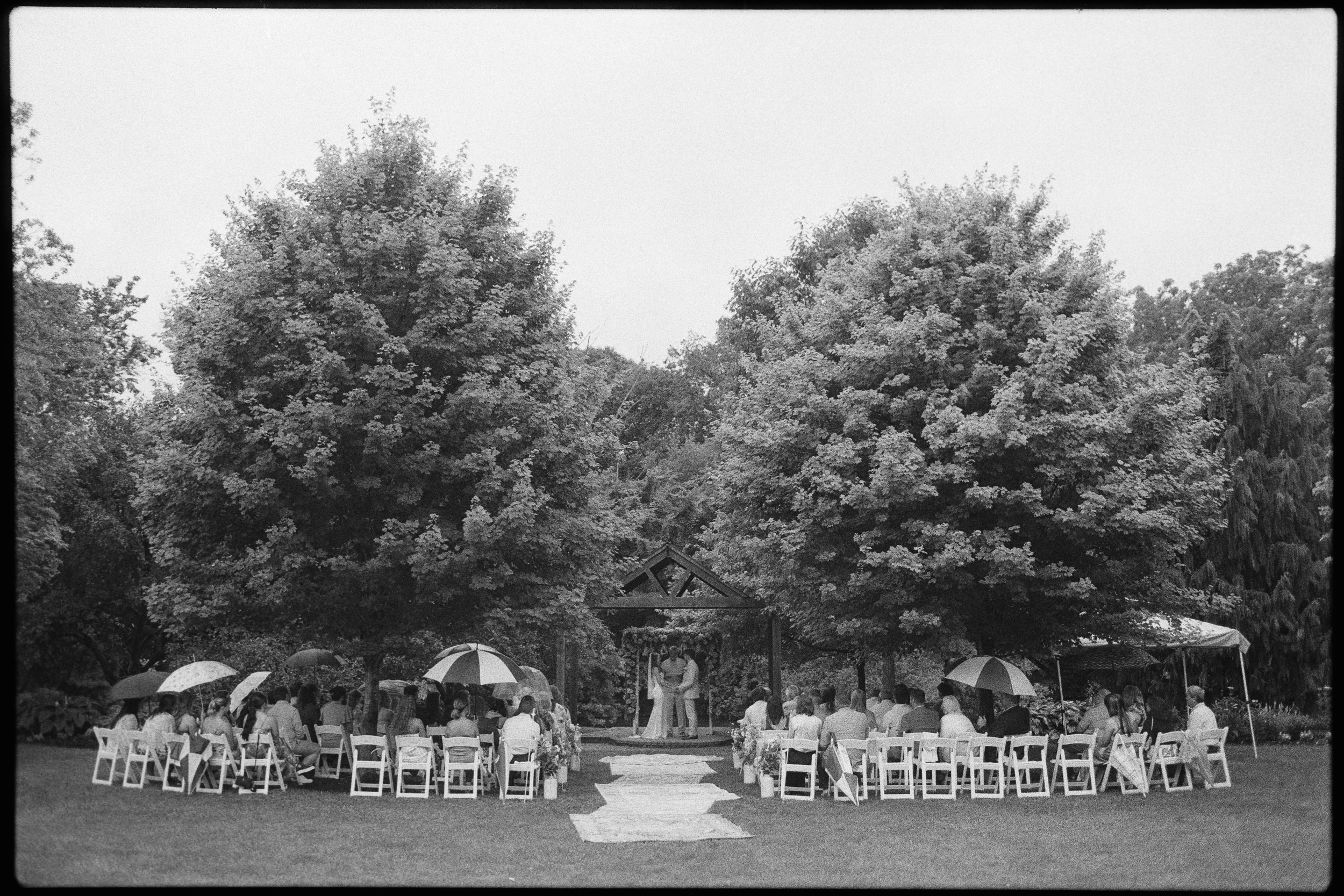 Black and white photo of an outdoor gathering with people sitting on chairs and holding umbrellas, beneath large trees, with a small stage or platform in the center.