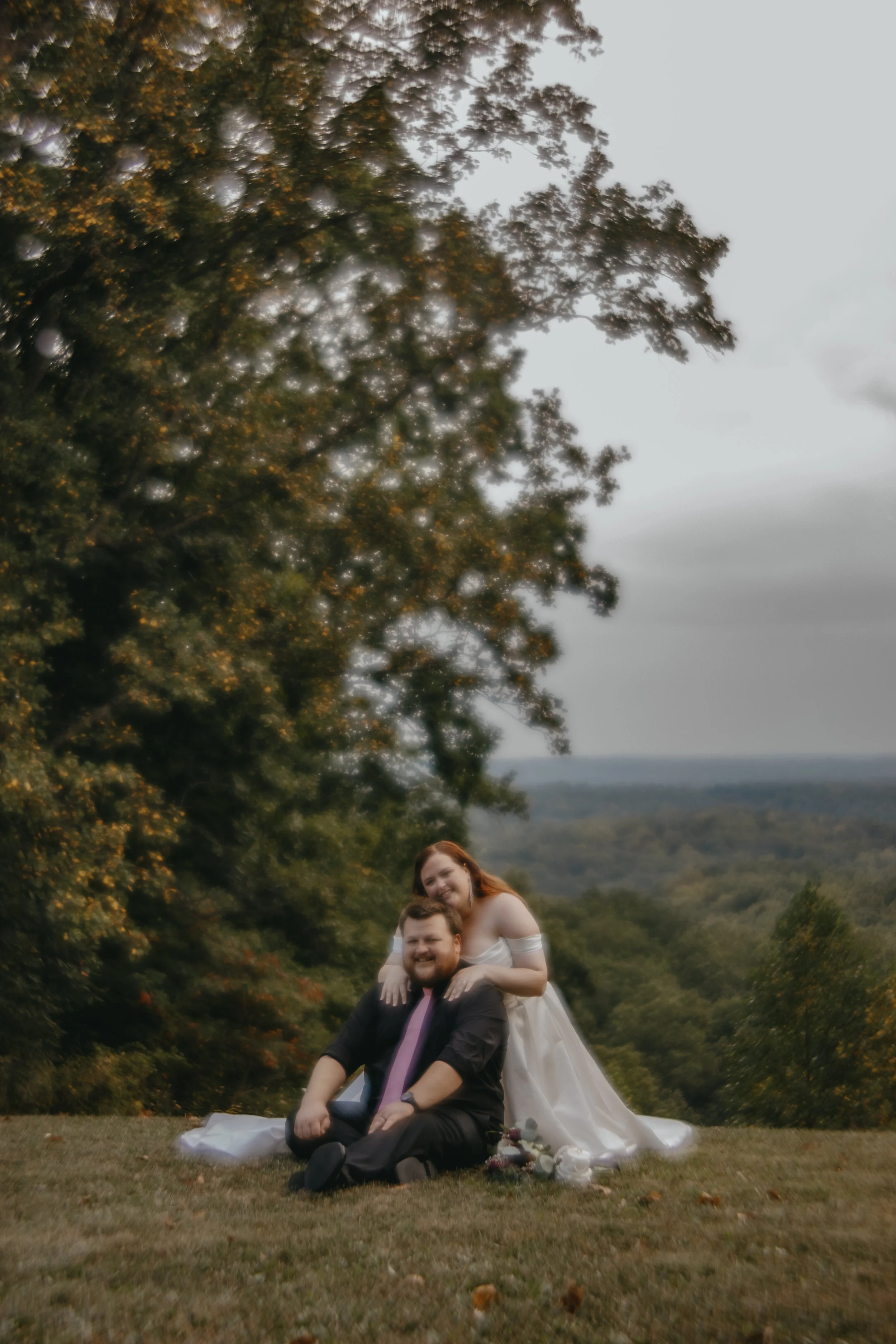 A couple in wedding attire sitting on grass with trees and cloudy sky in the background. The bride is on the groom's back, smiling, with flowers nearby.