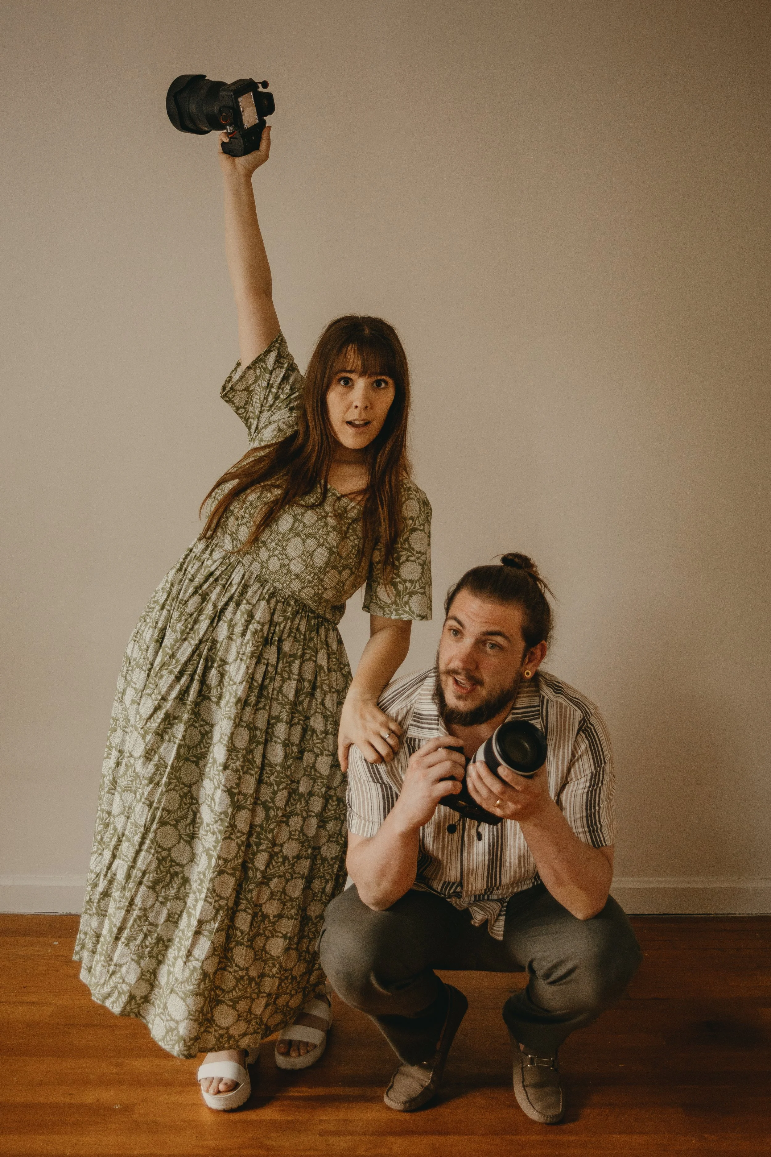 people are posing for a photo indoors. The woman with long brown hair, wearing a patterned dress, stands with her arm raised holding a camera. The man with a beard, wearing a striped shirt, glasses, and dark pants, is crouched in front holding a camera.