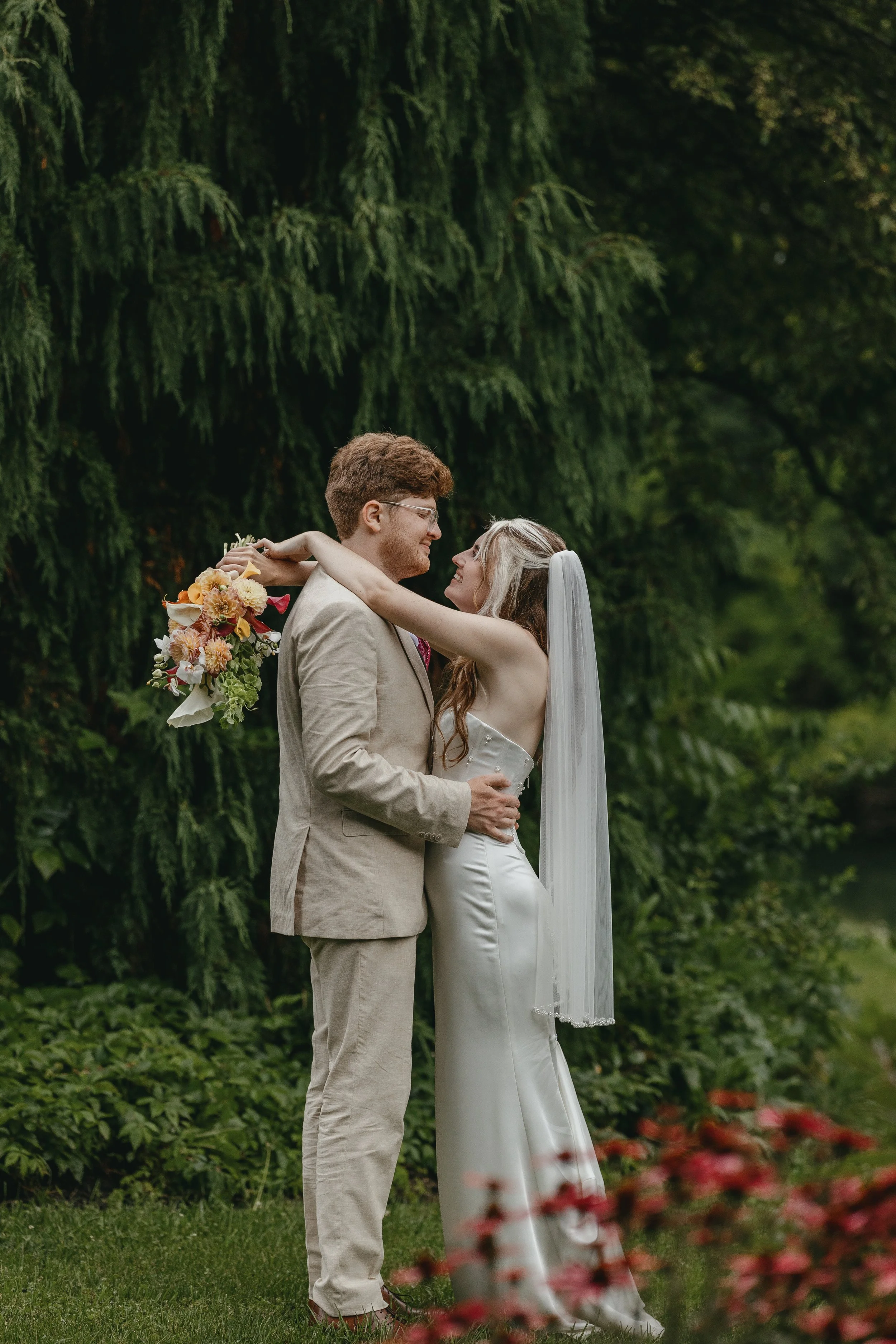 A bride and groom embrace in an outdoor setting with lush green trees in the background. The bride is holding a bouquet of flowers and has her arms around the groom, both smiling at each other.