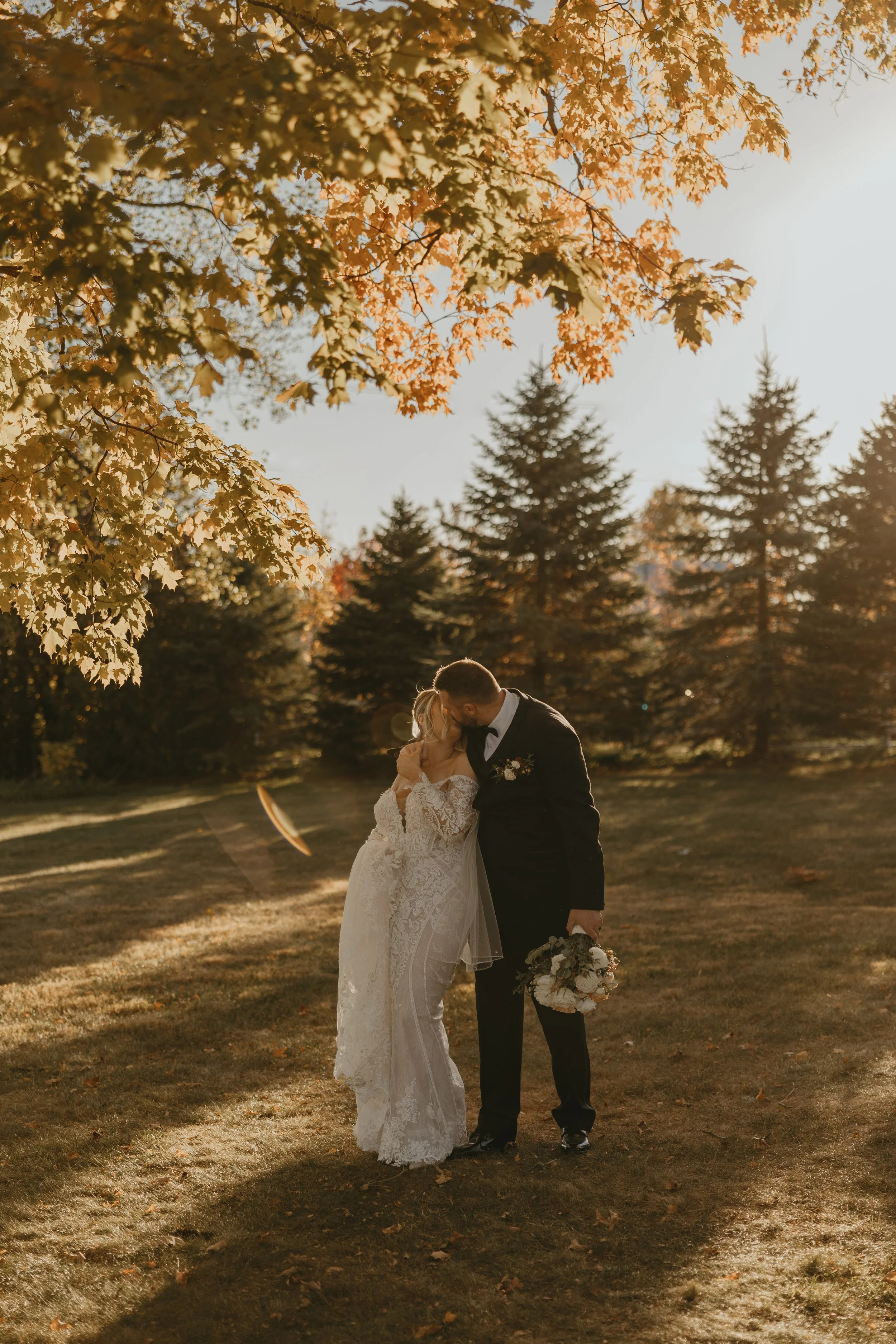 A bride and groom sharing a kiss outdoors during sunset, surrounded by autumn trees with orange leaves, holding a bouquet of flowers.