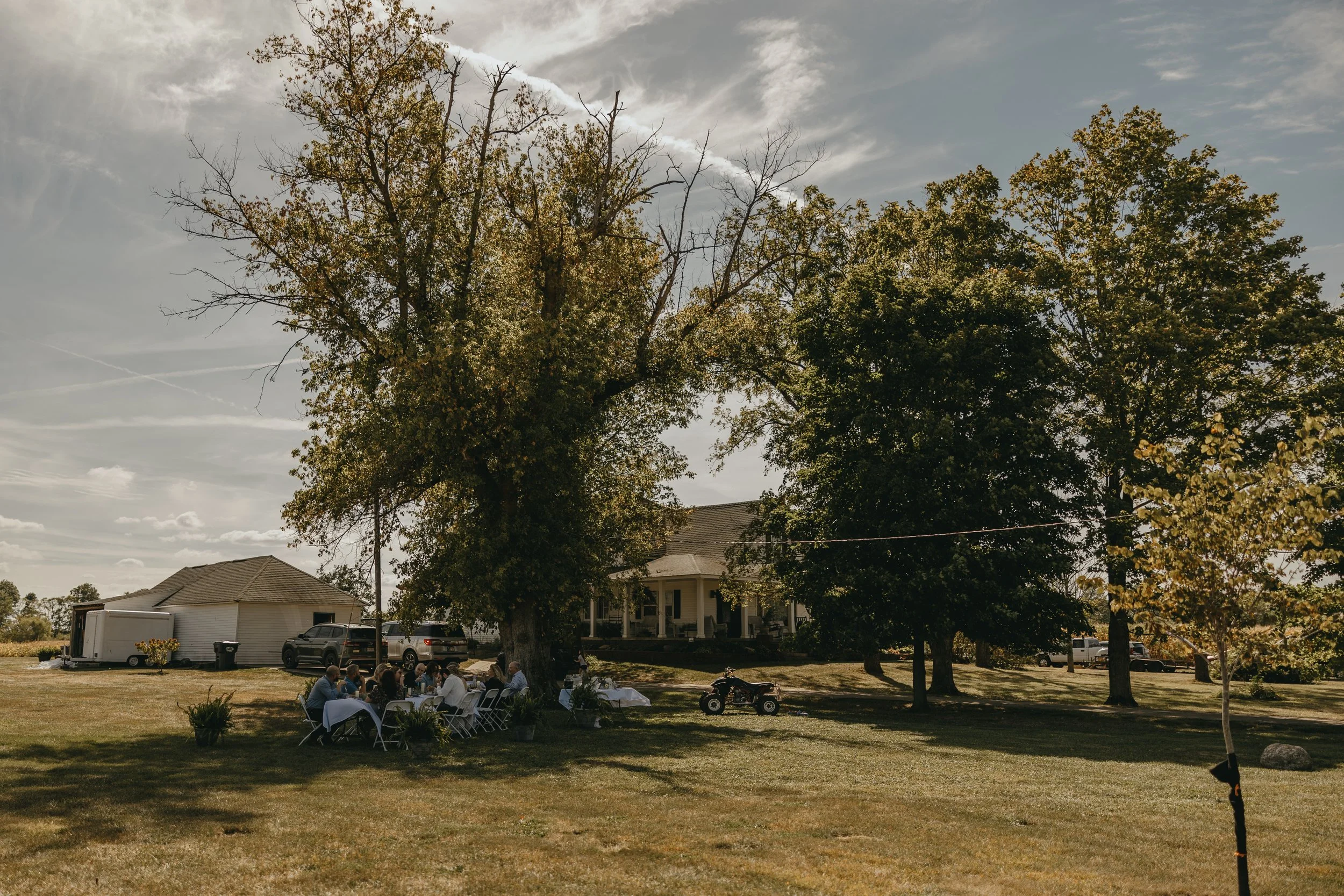 People sitting at tables outdoors in a grassy yard near large trees, with a house and a garage in the background on a sunny day. Shot by Wedding Photographers Dunckel Haus Photography