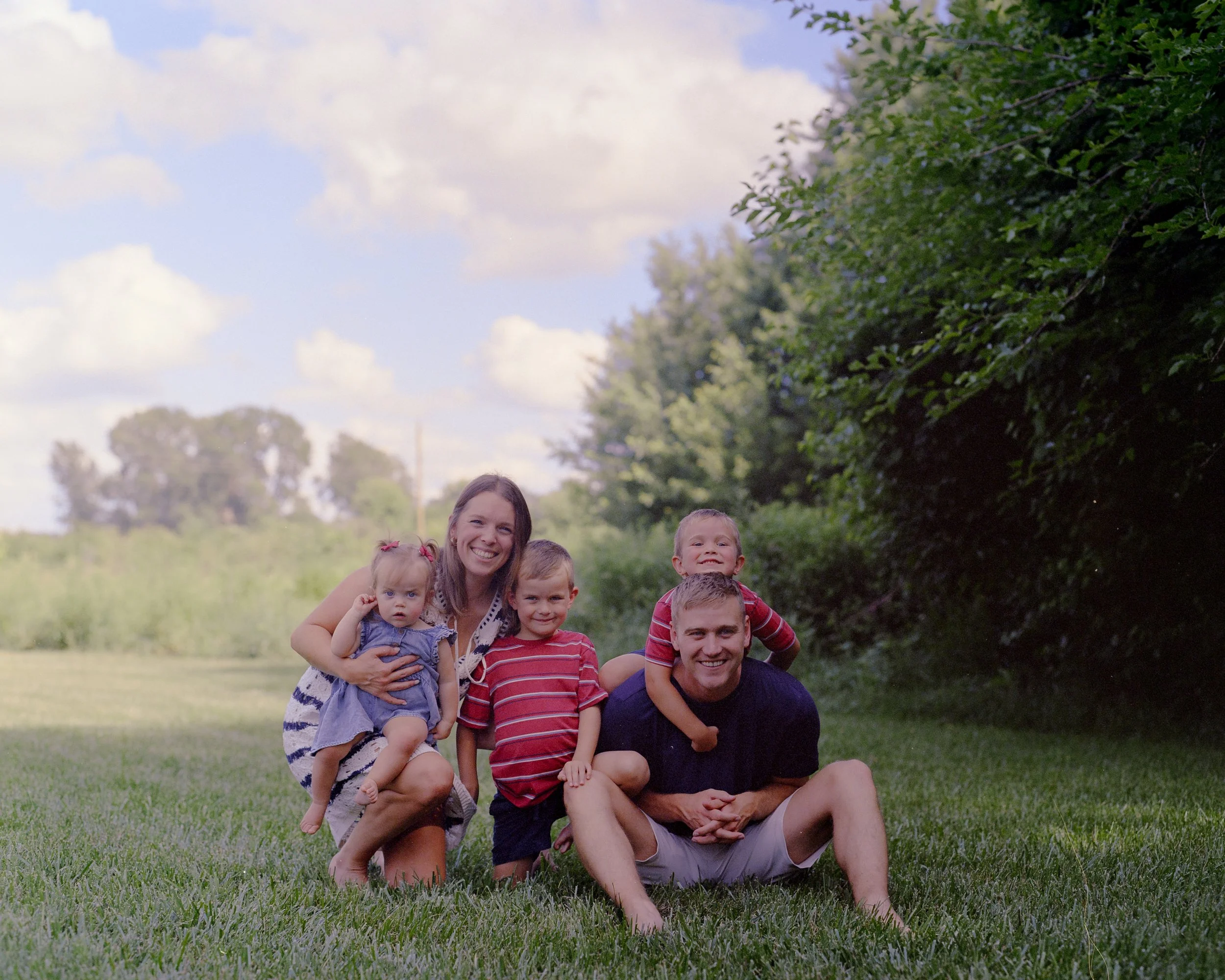 A happy family of seven, including a woman, a man, and five children, posing outdoors on a grassy field with trees and a cloudy sky in the background.