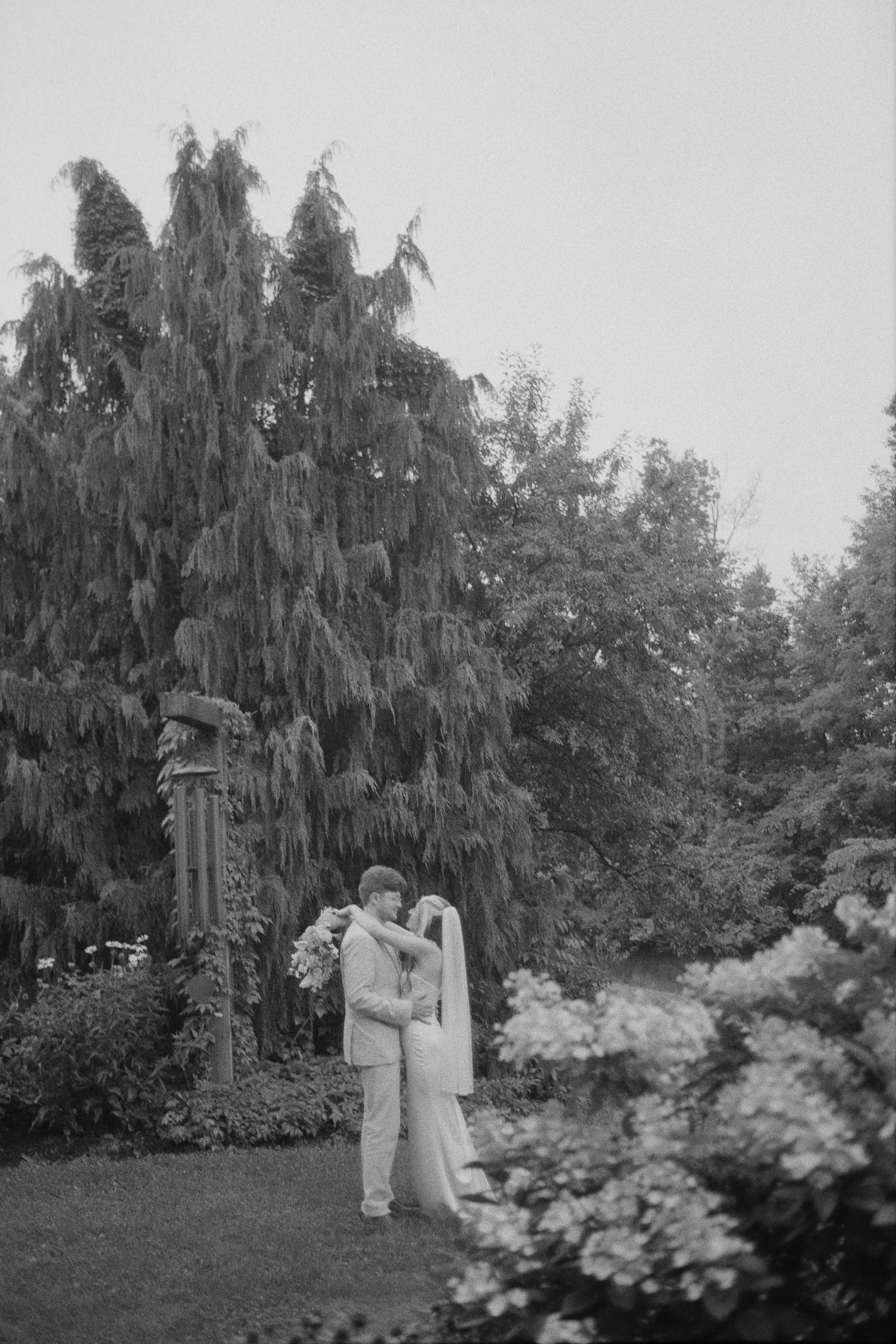 A couple standing close together in an outdoor setting surrounded by trees and flowers, with the man holding the woman's hands.