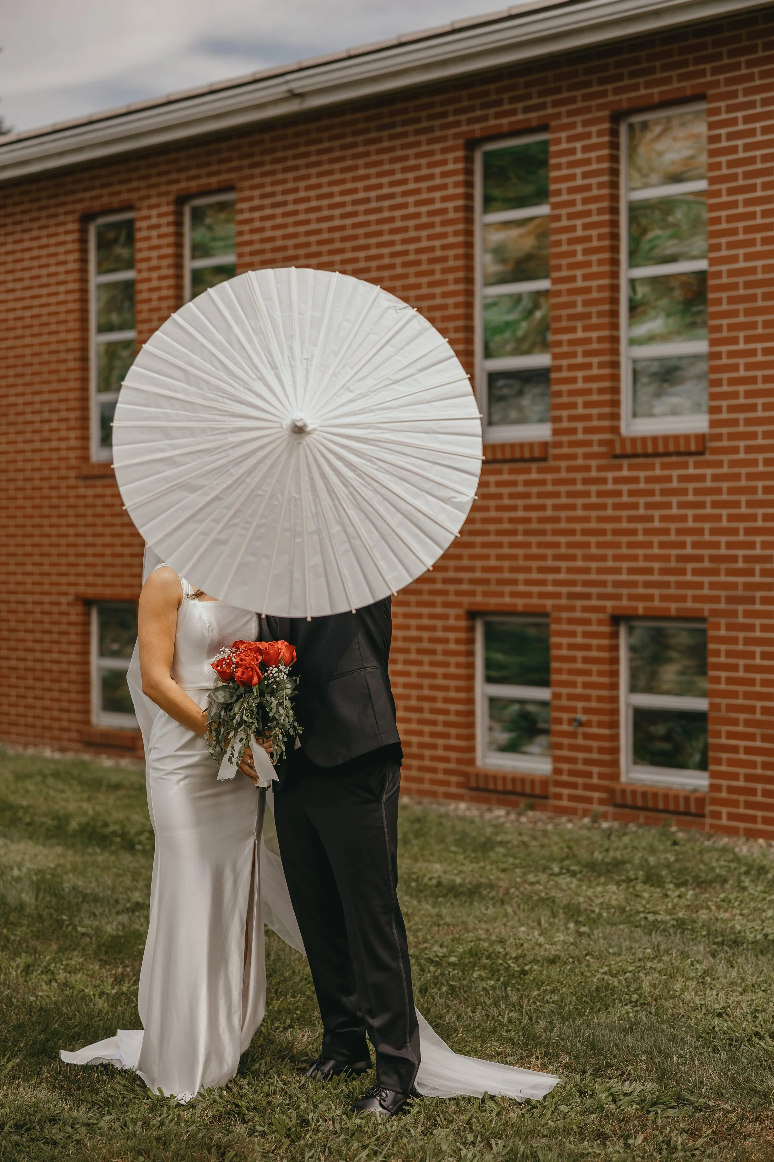 A bride and groom holding a white parasol with a brick building in the background. The bride is holding a bouquet of red and white flowers. The couple is standing on grass, and the bride is wearing a white wedding dress, while the groom is in a black