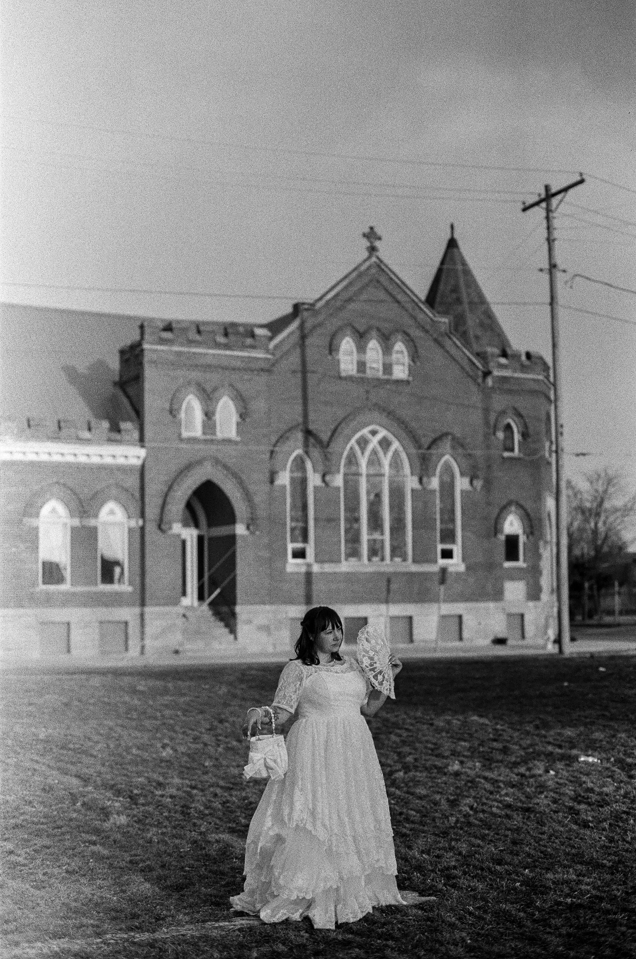 A woman in a vintage wedding dress holding a bouquet and a paper bag stands outdoors in front of a large Victorian-style church building with stained glass windows and a peaked roof.