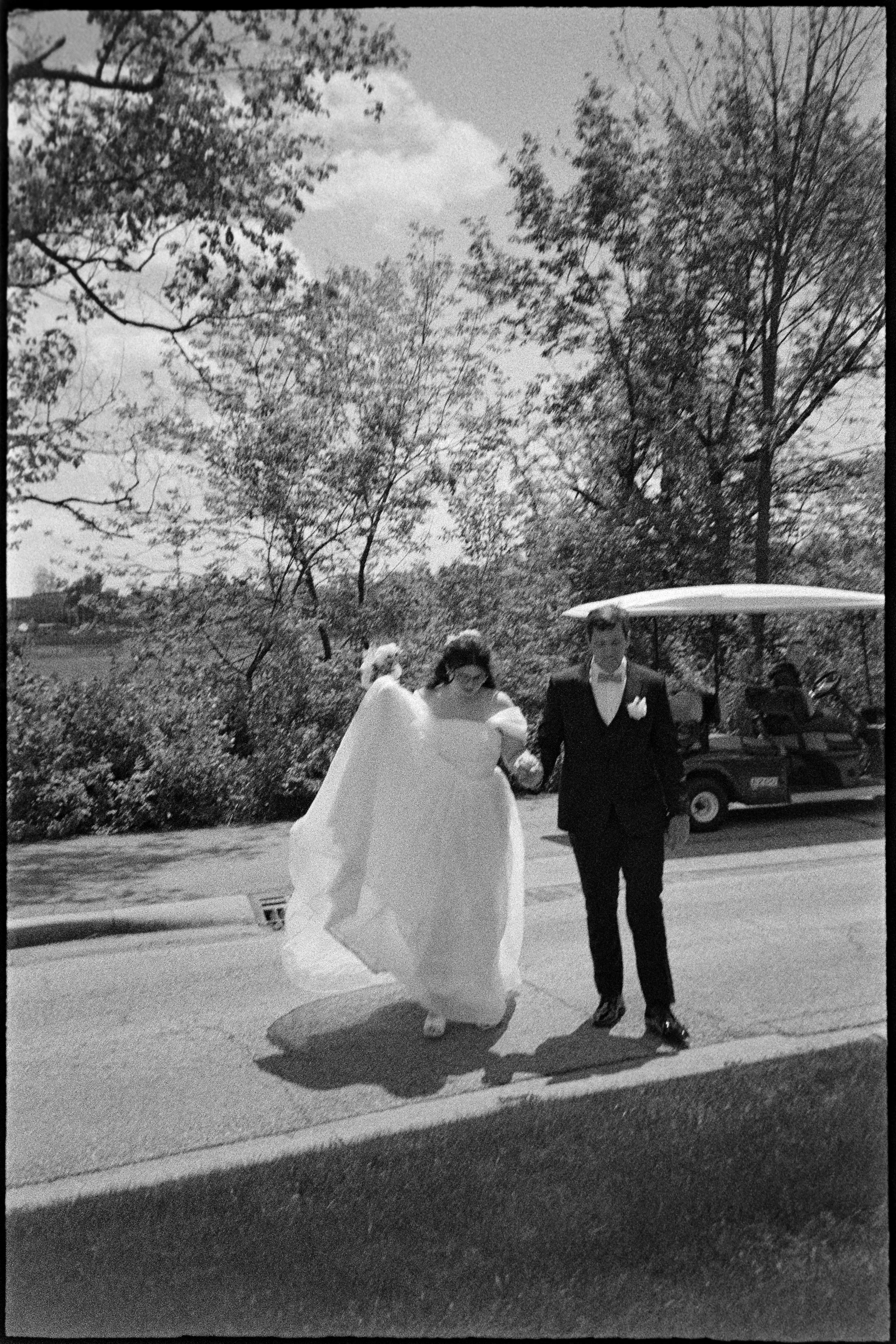 A bride and groom walking together outdoors near a golf cart, with trees and bushes in the background, on a sunny day.