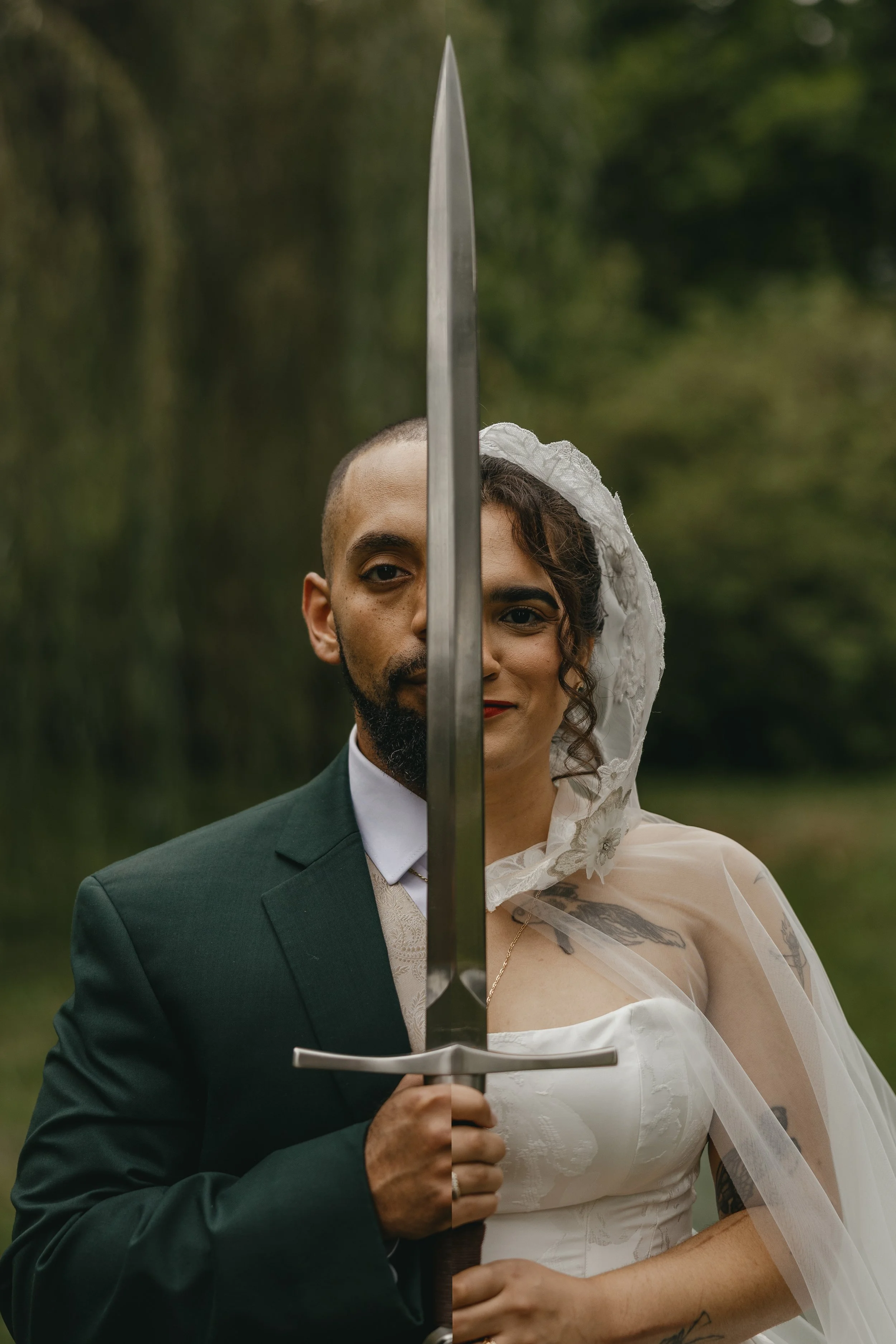 A man in a green suit and a woman in a white dress with a lace bonnet holding a sword together, an outdoor setting with green trees in the background.