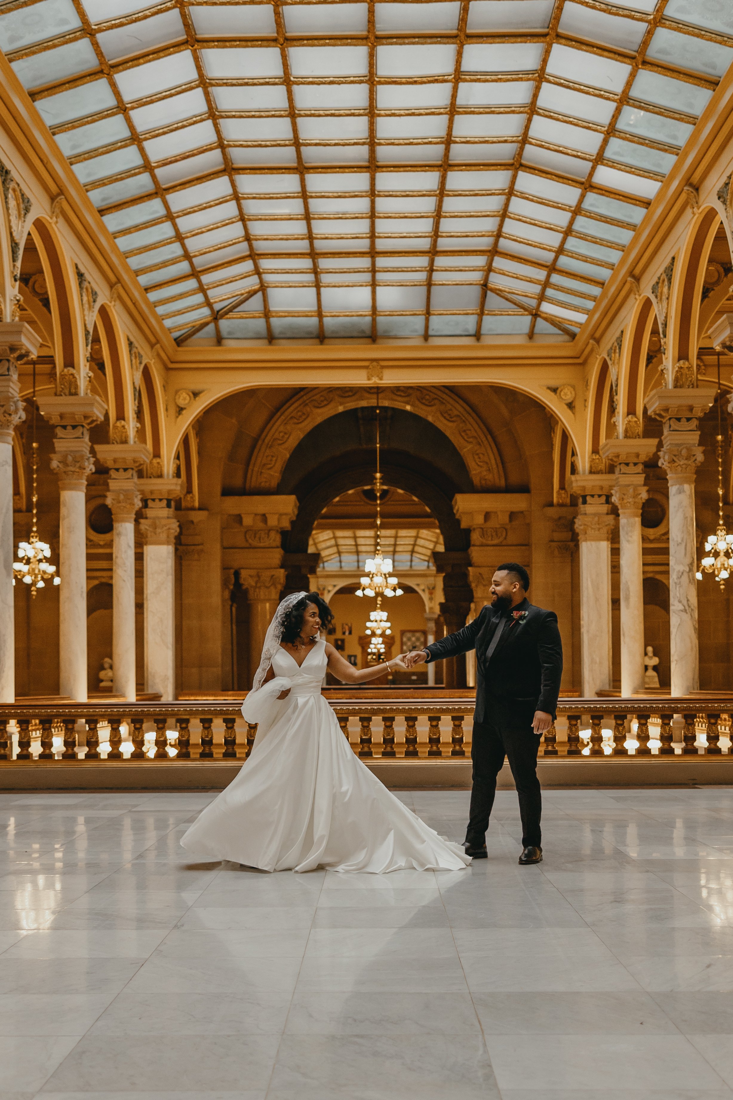 A bride and groom dancing in a grand, ornate hall with a glass ceiling and classical columns. Shot by Wedding Photographers Dunckel Haus Photography
