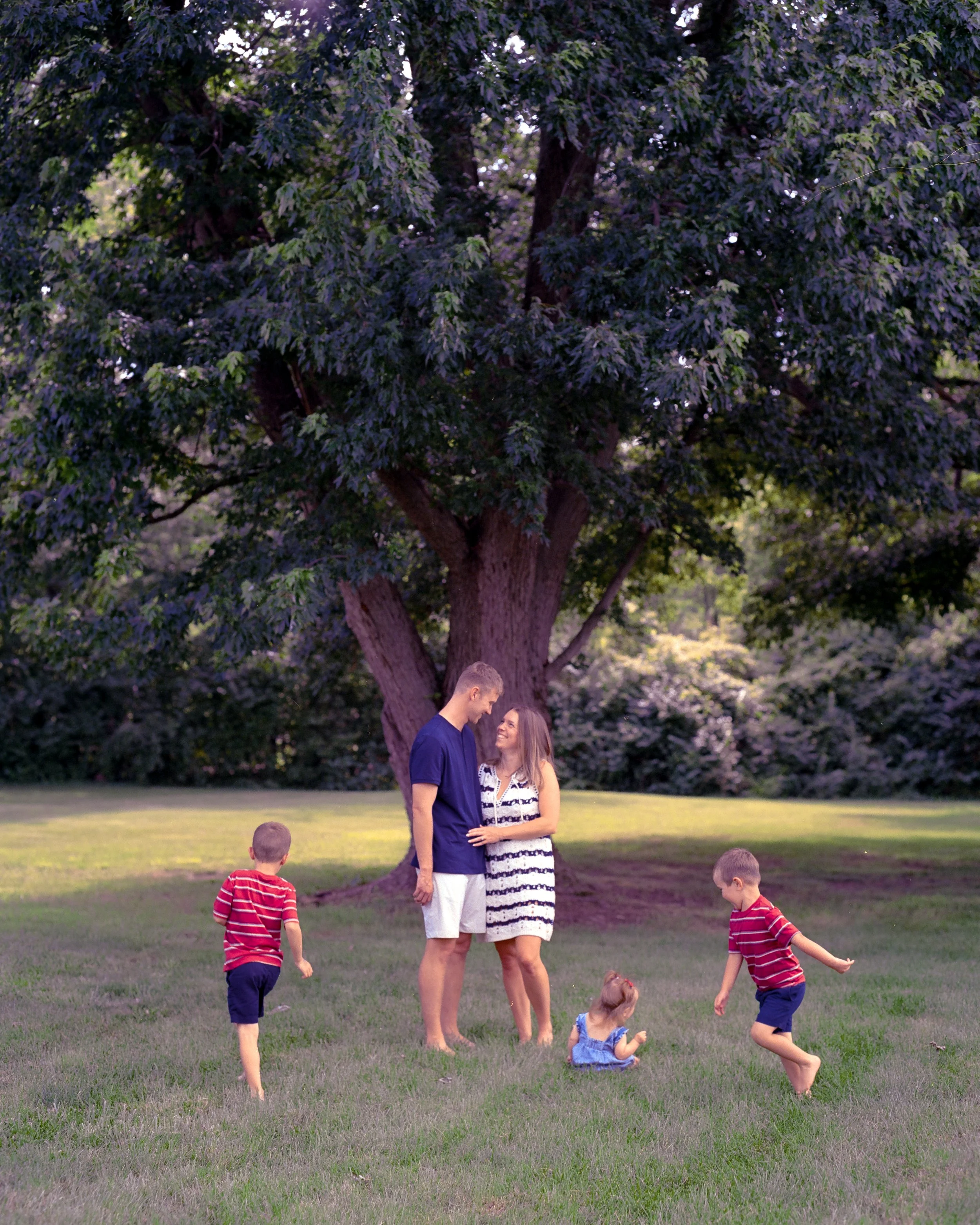 A family of five standing and playing on a grassy lawn in front of a large tree with dense green leaves. The two adults, a man and a woman, are standing close together, and three children are playing nearby.