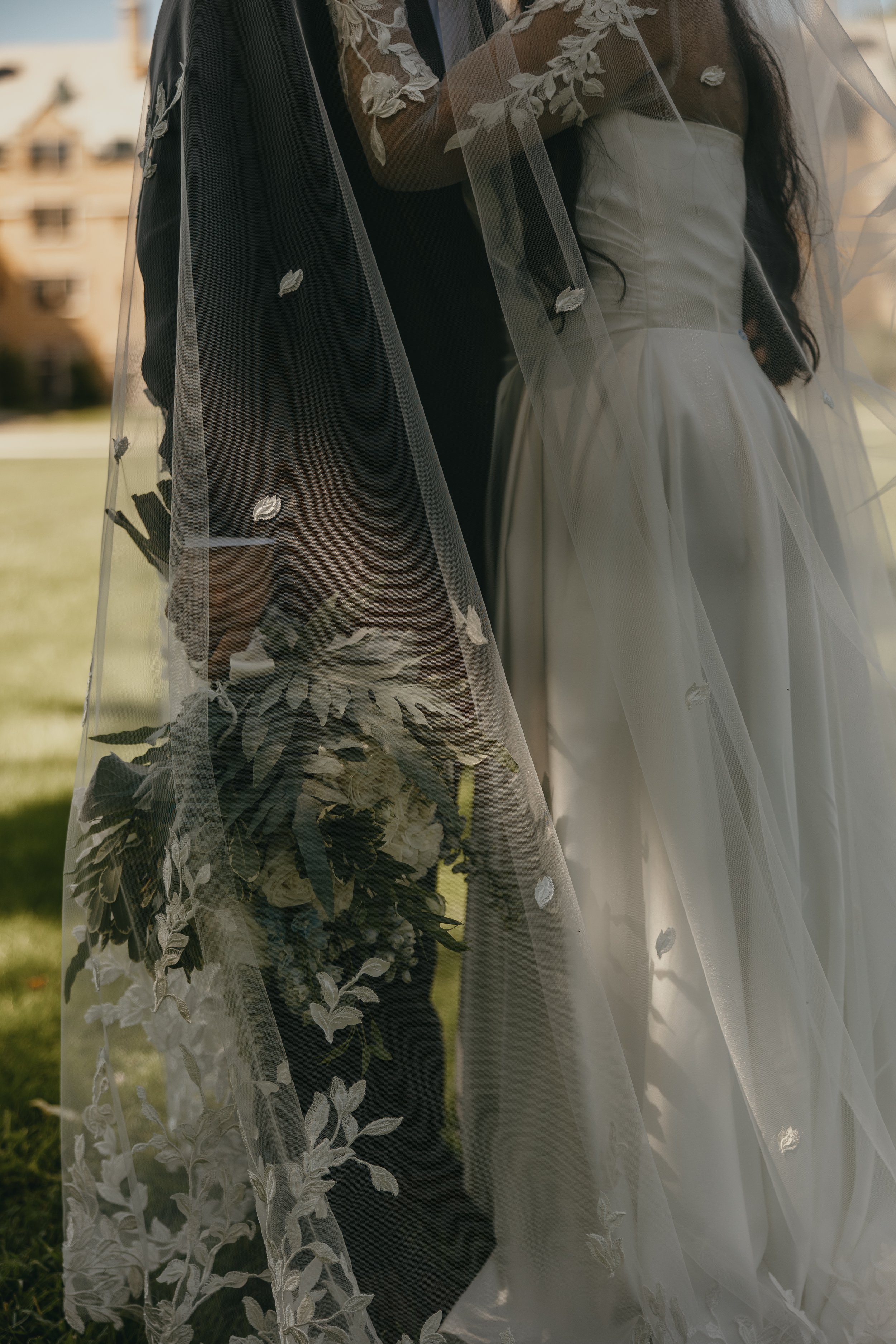 Close-up of a bride in a white wedding dress and a groom in a black suit, behind a sheer veil with floral embroidery, with a building and grass in the blurry background.