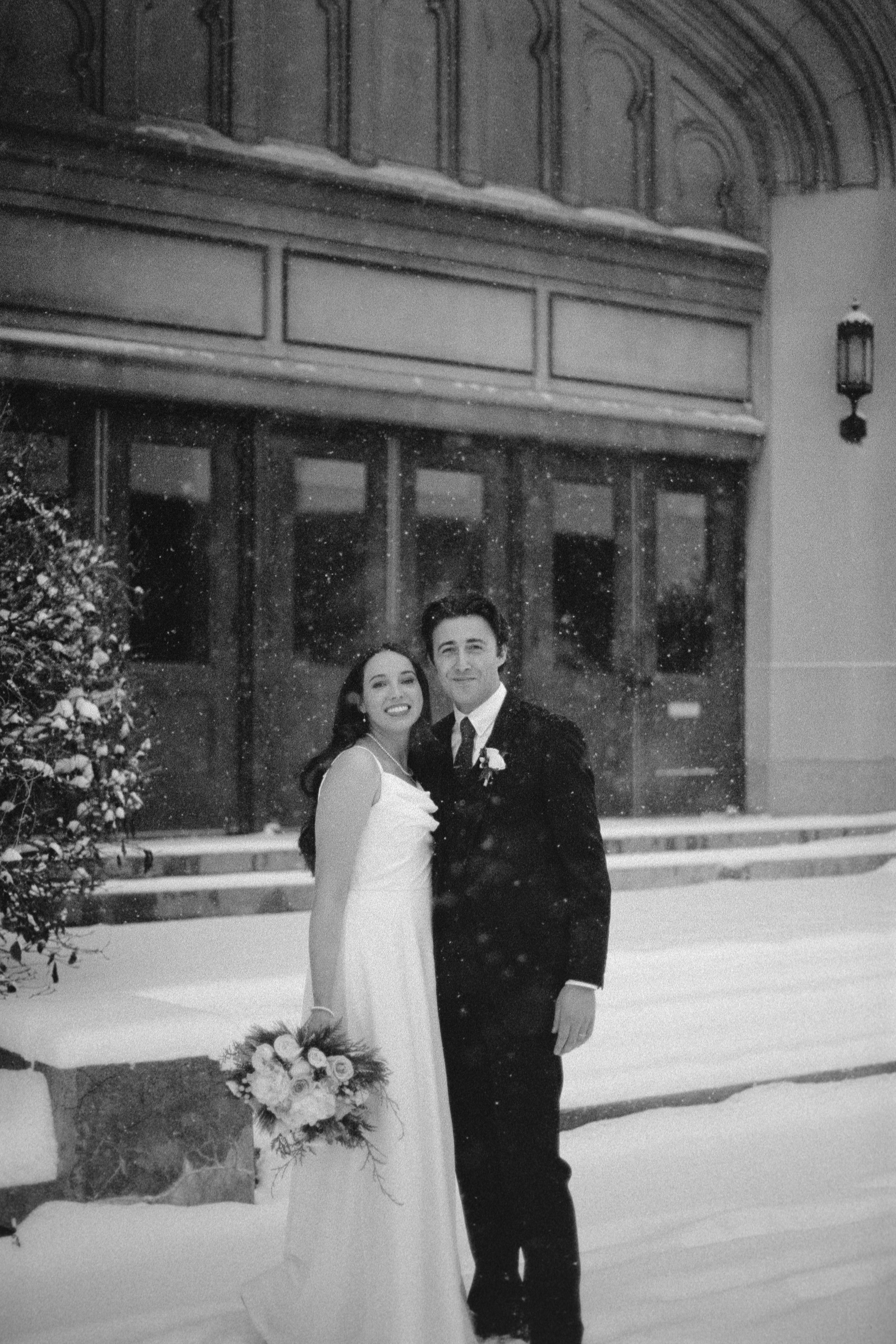A wedding couple standing outside in snow in front of a building with large windows and a balcony. The bride is wearing a white wedding dress, and the groom is in a black suit. A bouquet of flowers is on the ground next to them.