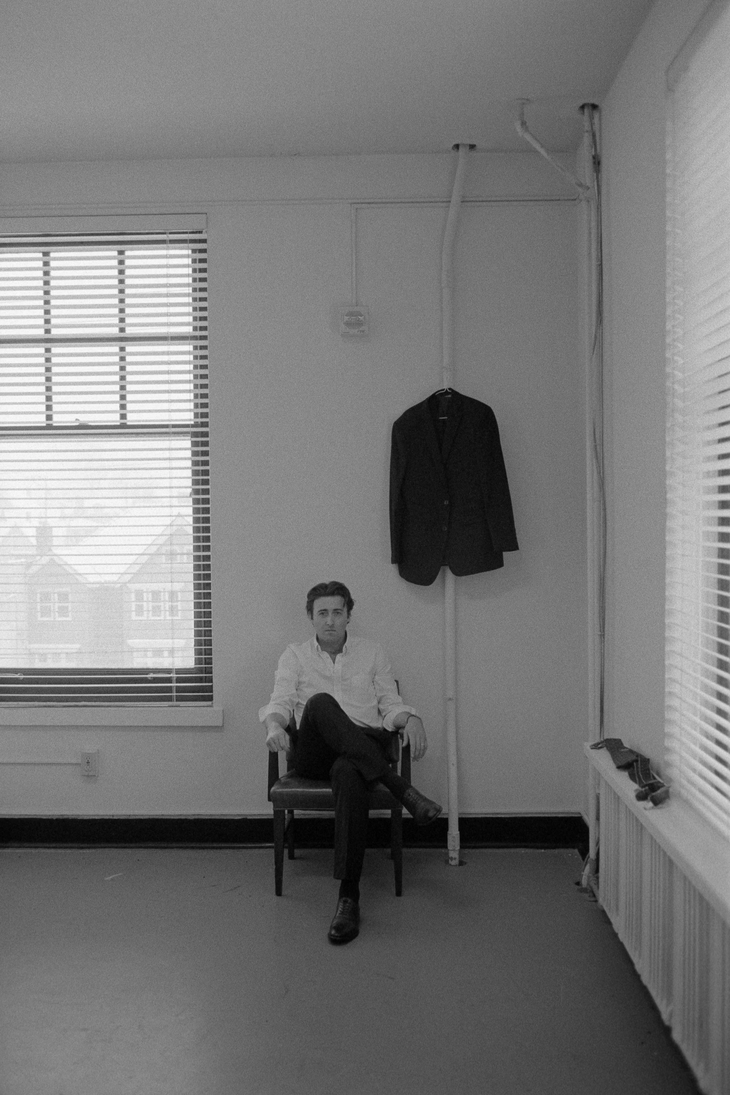 A man wearing a white shirt and black pants, sitting on a chair with crossed legs, in an office-like room with large windows, partially blinds, and a coat hanging on the wall.