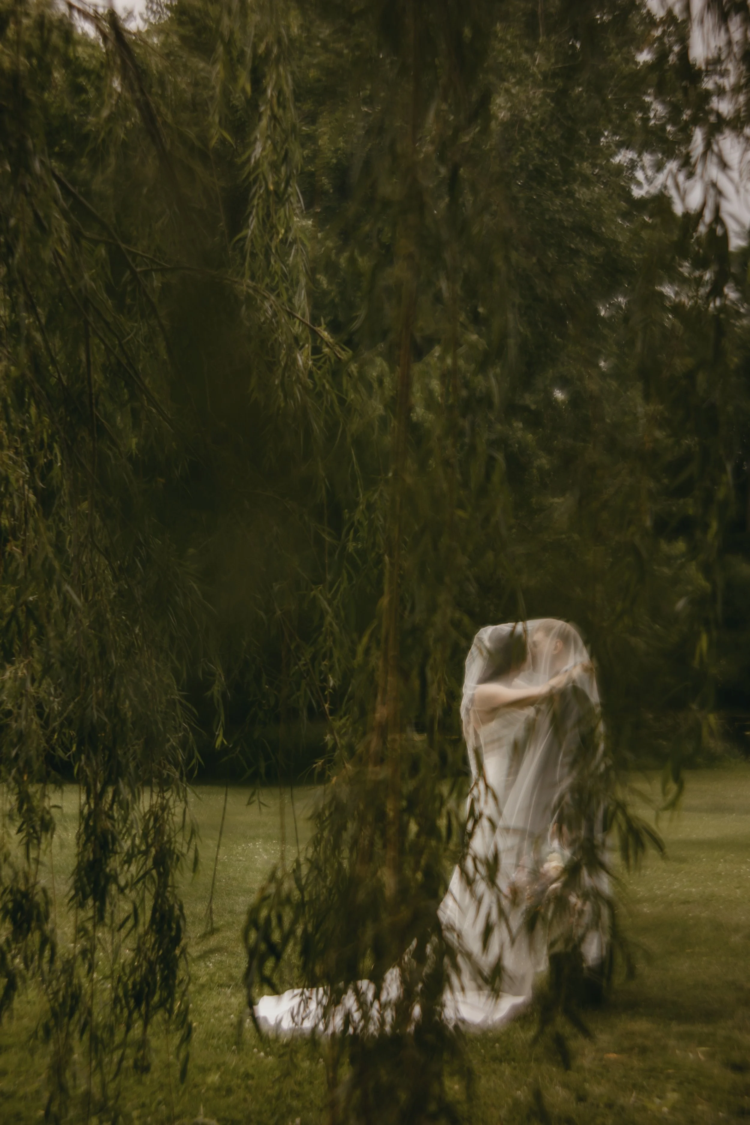 A couple dressed in white wedding garments standing on a grassy area outdoors, seen through the hanging branches of a tree, with a calm body of water in the background.