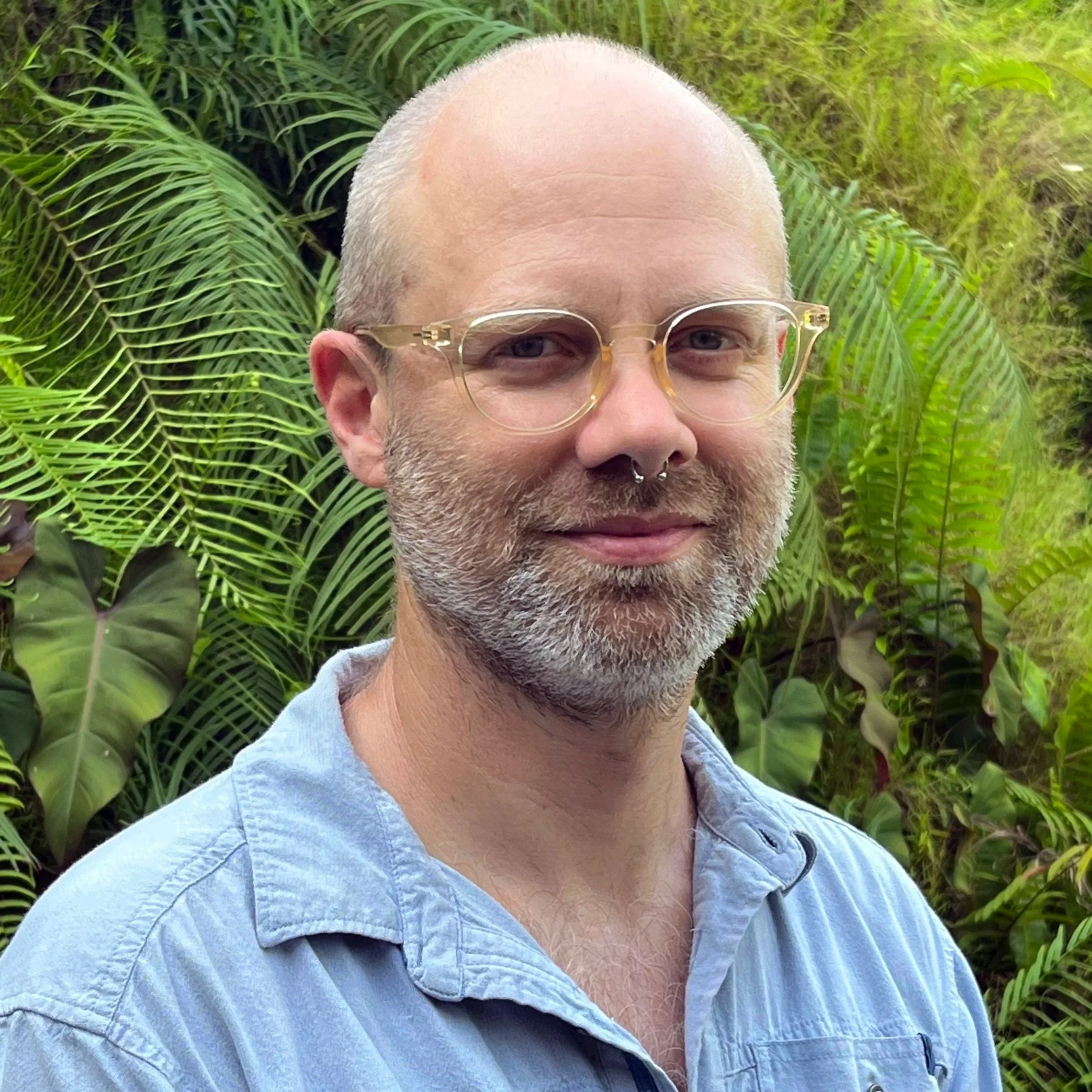 A man with glasses and a short beard standing outdoors in front of purple and green foliage, smiling at the camera.