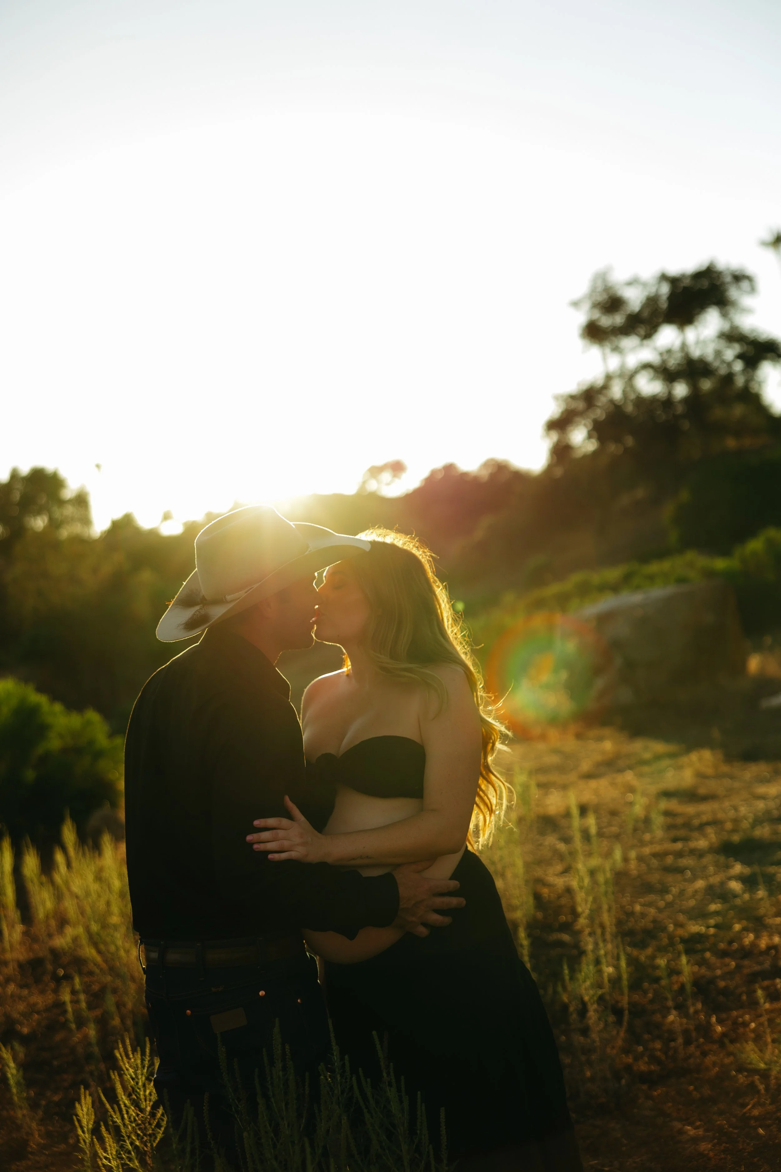 A man and woman kissing outdoors at sunset, with the man wearing a cowboy hat and the woman in a strapless black top. The sun is setting behind them, creating a warm glow.