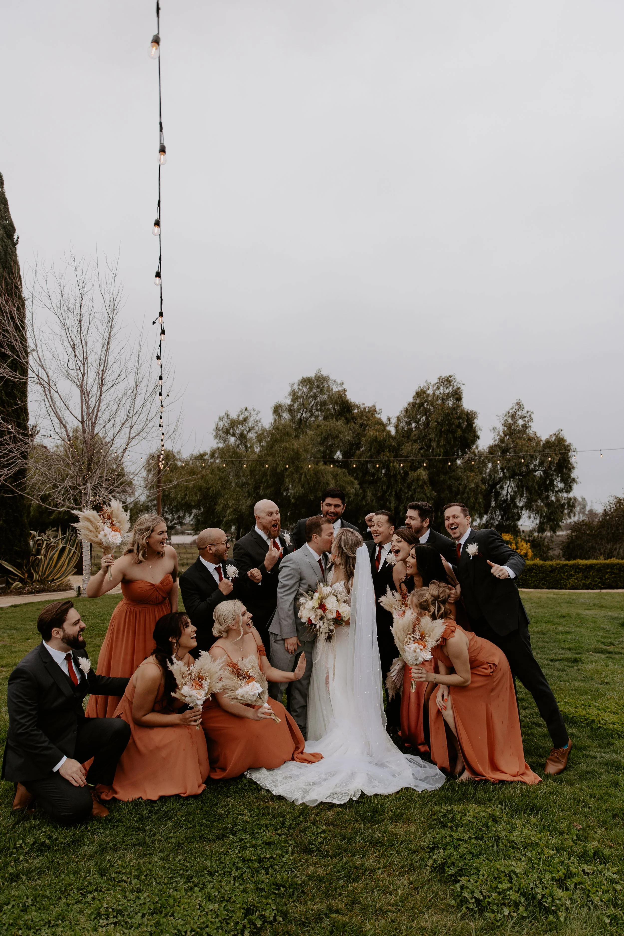 A wedding party outdoors with the bride in a white dress and veil, surrounded by bridesmaids in rust-colored dresses and groomsmen in black suits, gathered on a grassy area with trees and string lights overhead.