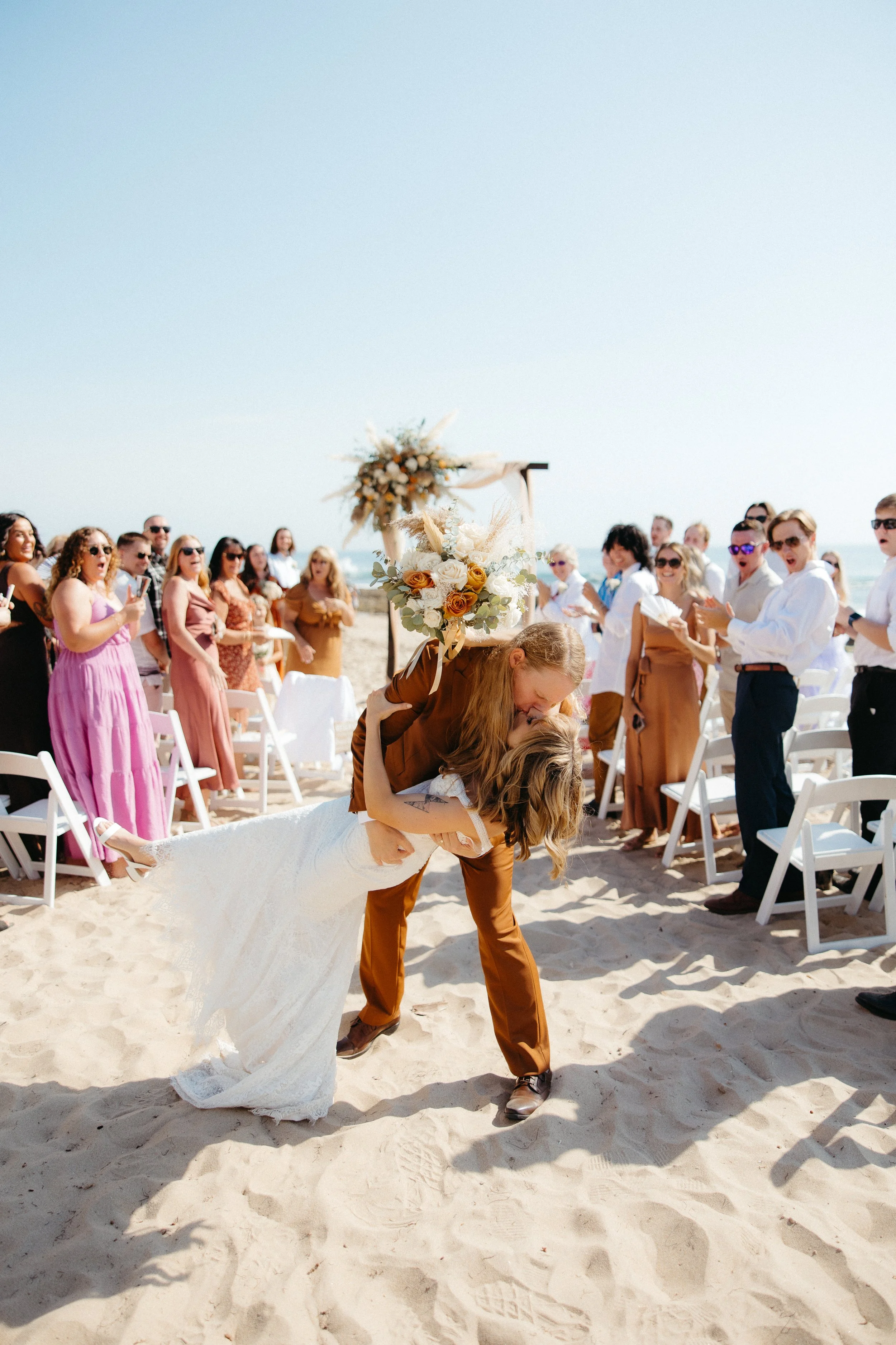 A couple kissing at a beach wedding, with guests clapping and taking photos in the background, and a floral wedding arch behind them.