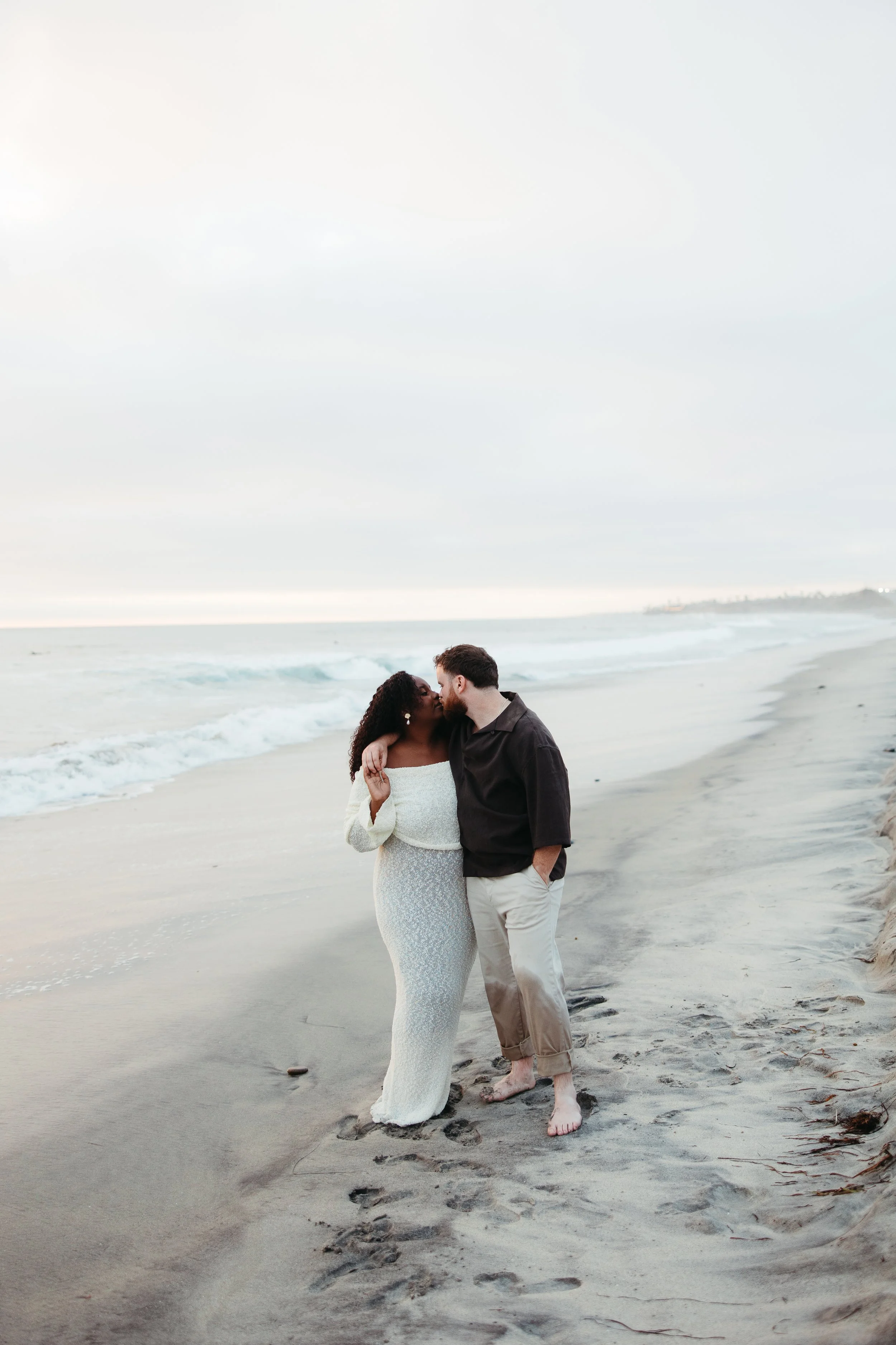 A couple shares a kiss on the beach at sunset, with the ocean waves behind them and footprints in the sand.