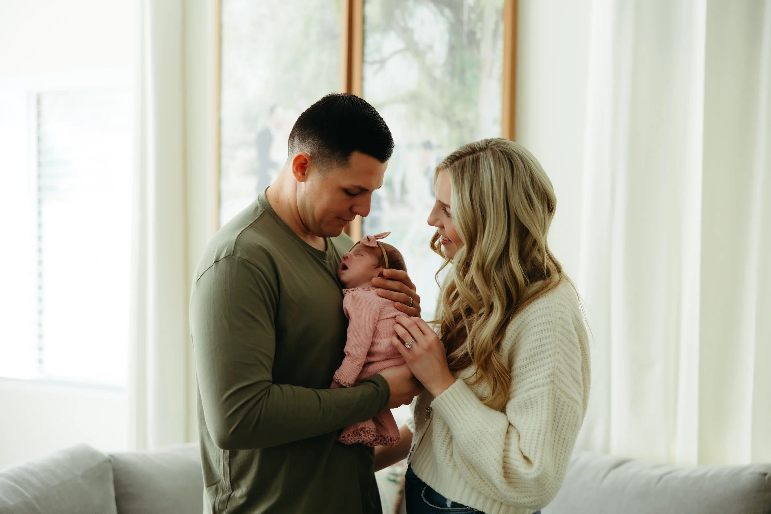 A young couple holding their newborn baby inside a bright room with large windows and cream-colored curtains.