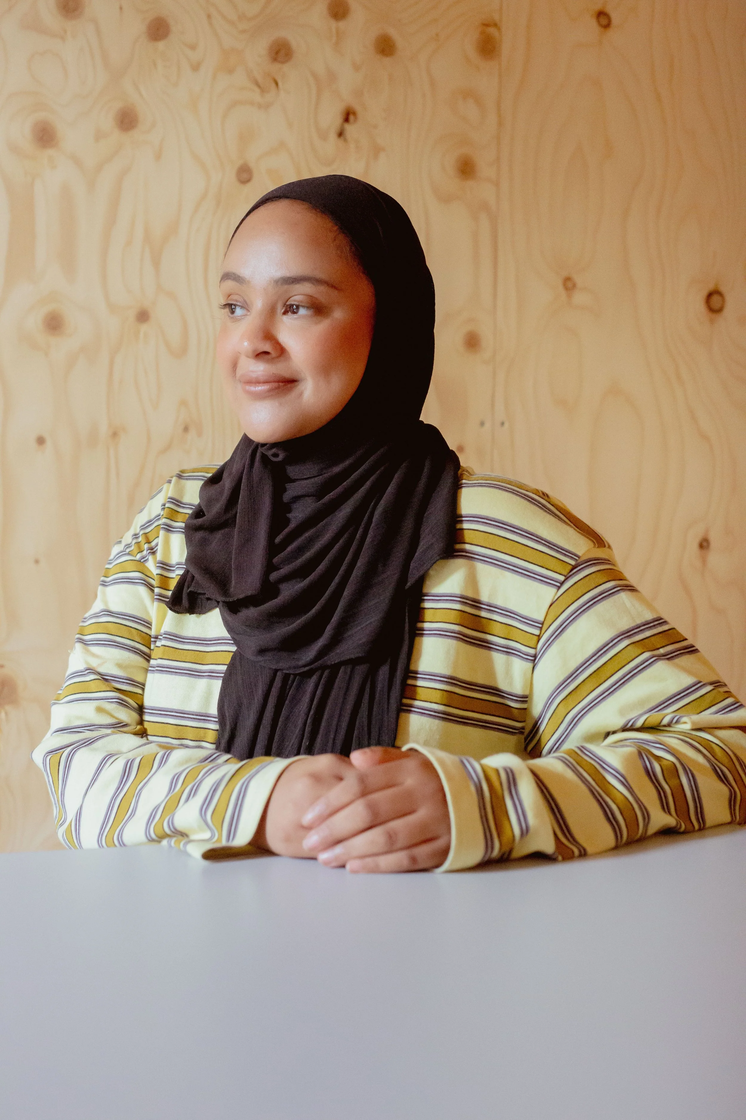 Fatima smiling warmly while seated at a table, wearing a black hijab and yellow striped sweater.