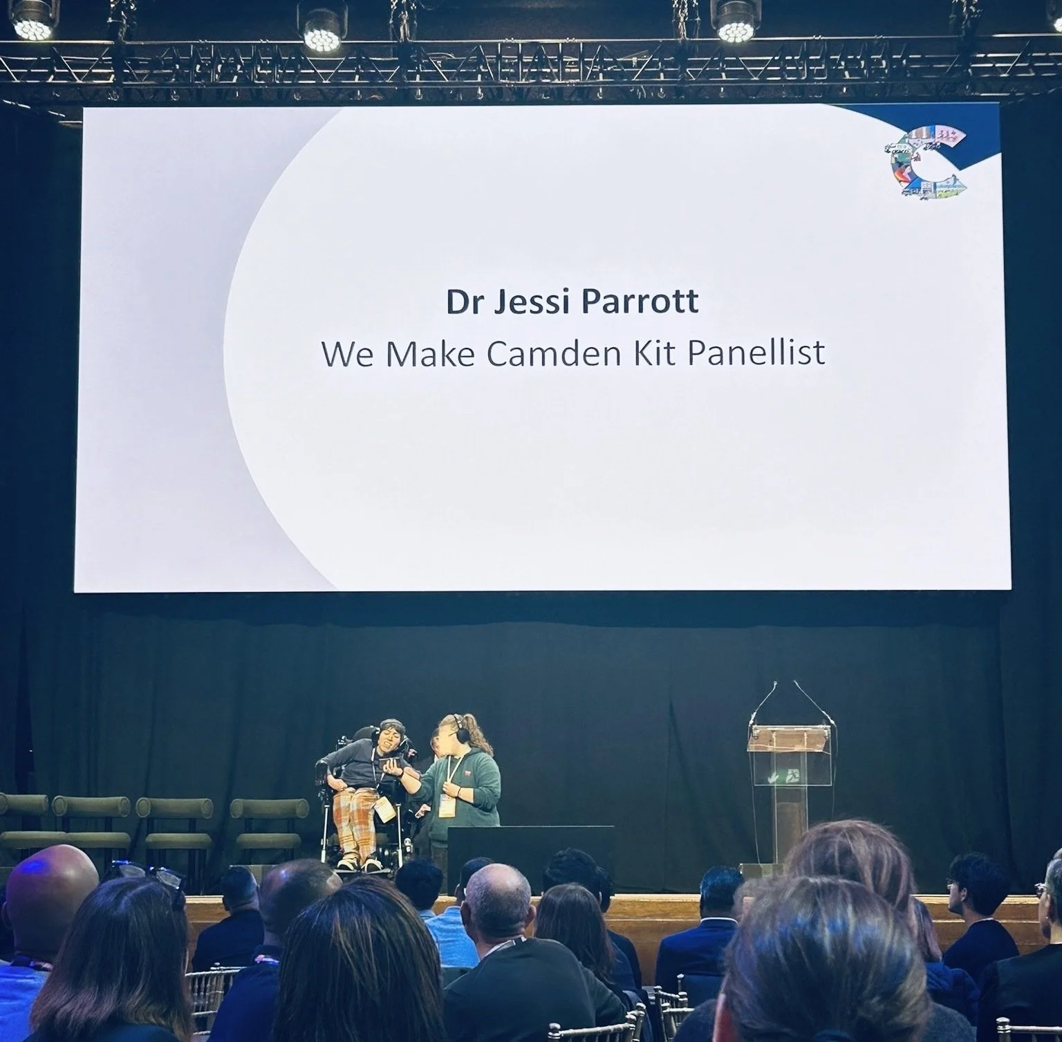 A stage where a person is giving a speech, behind them a screen says "Dr Jessi Parrott We Make Camden Kit Panellist".