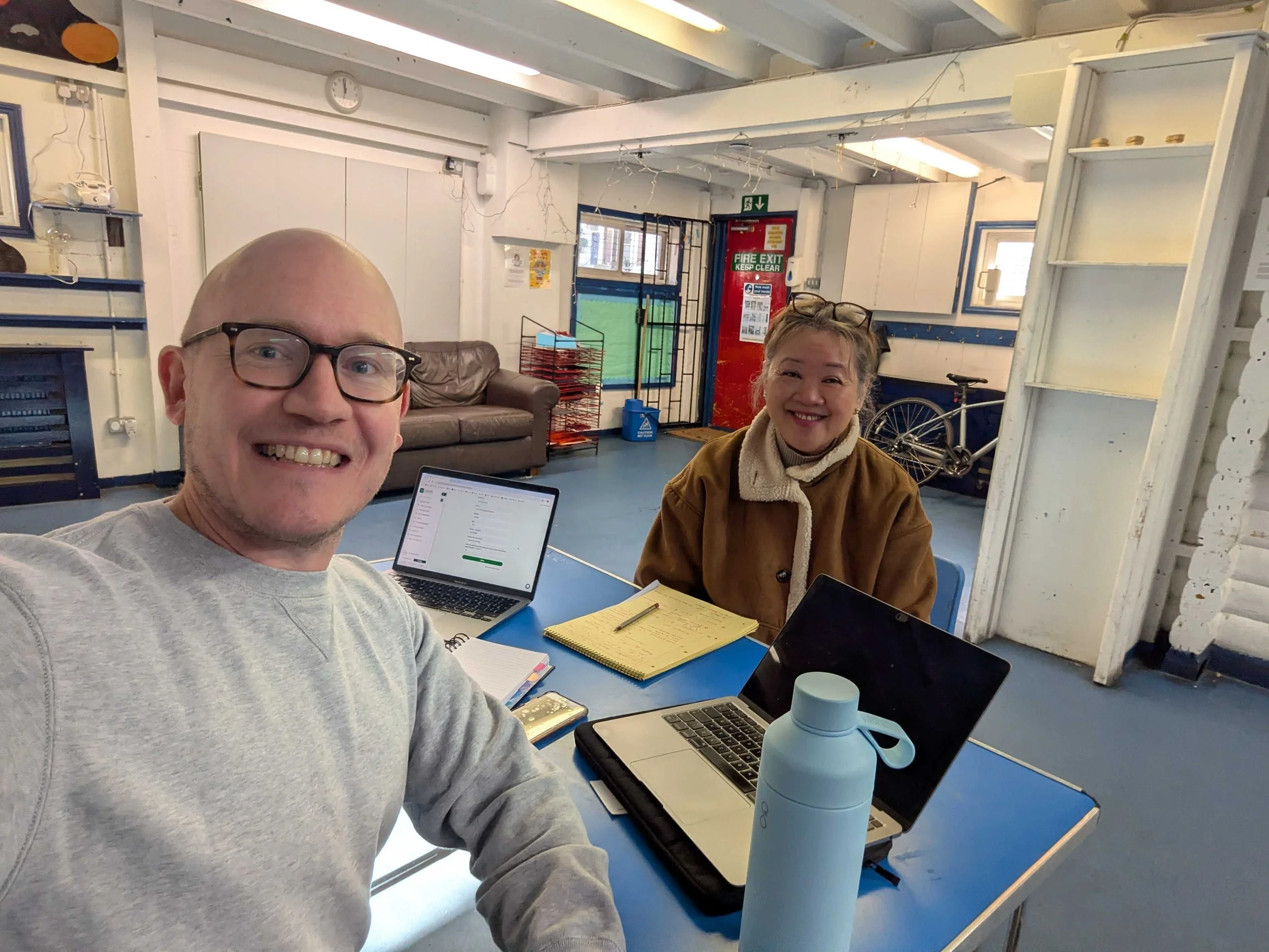 Grants Co-ordinator Andy Naylor smiling with an attendee at a Camden Giving grant drop-in session. They are sitting at a blue table with laptops and a notepad in a community centre.
