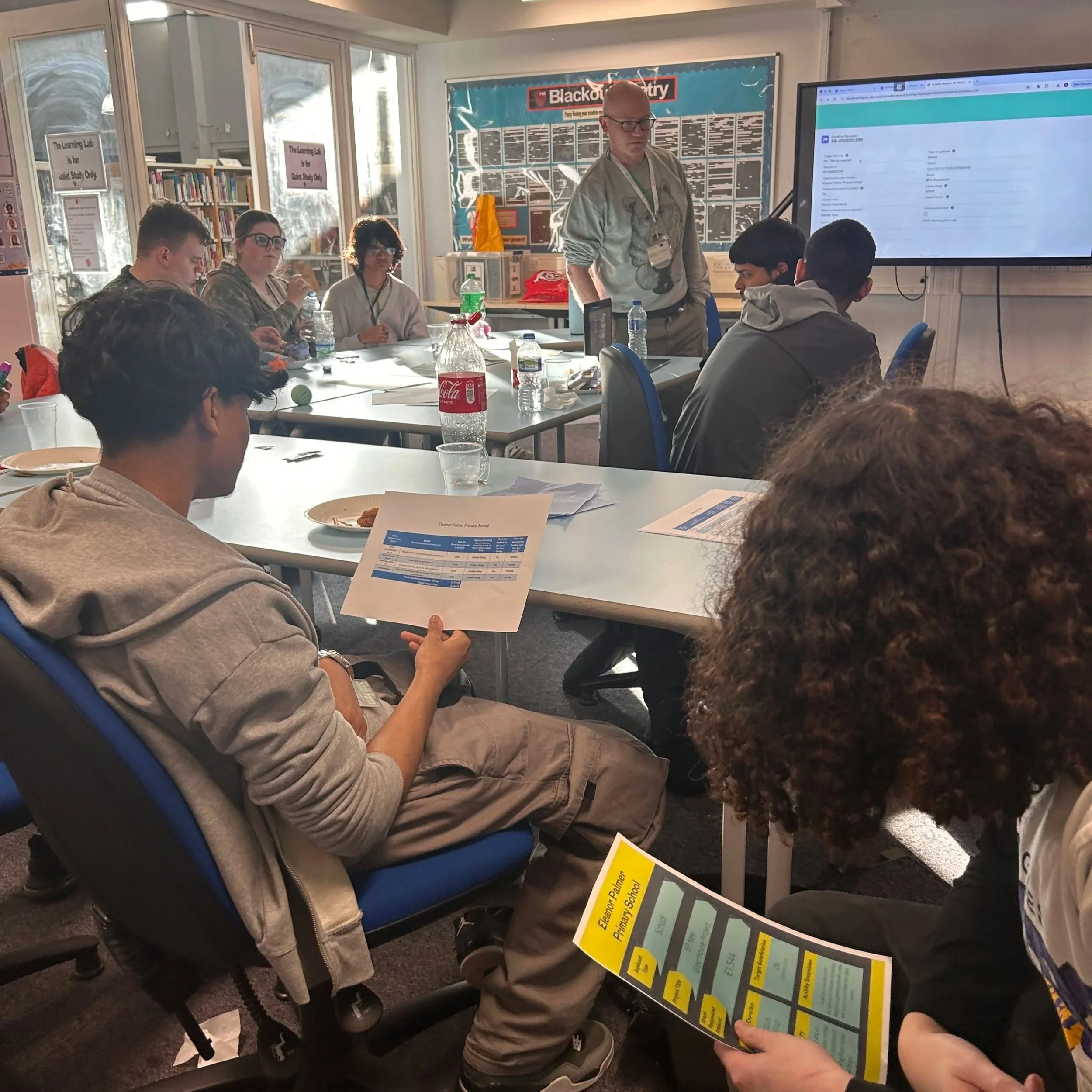 Young people sitting at a table review printed grant application documents and budget sheets while a facilitator stands nearby in a classroom.