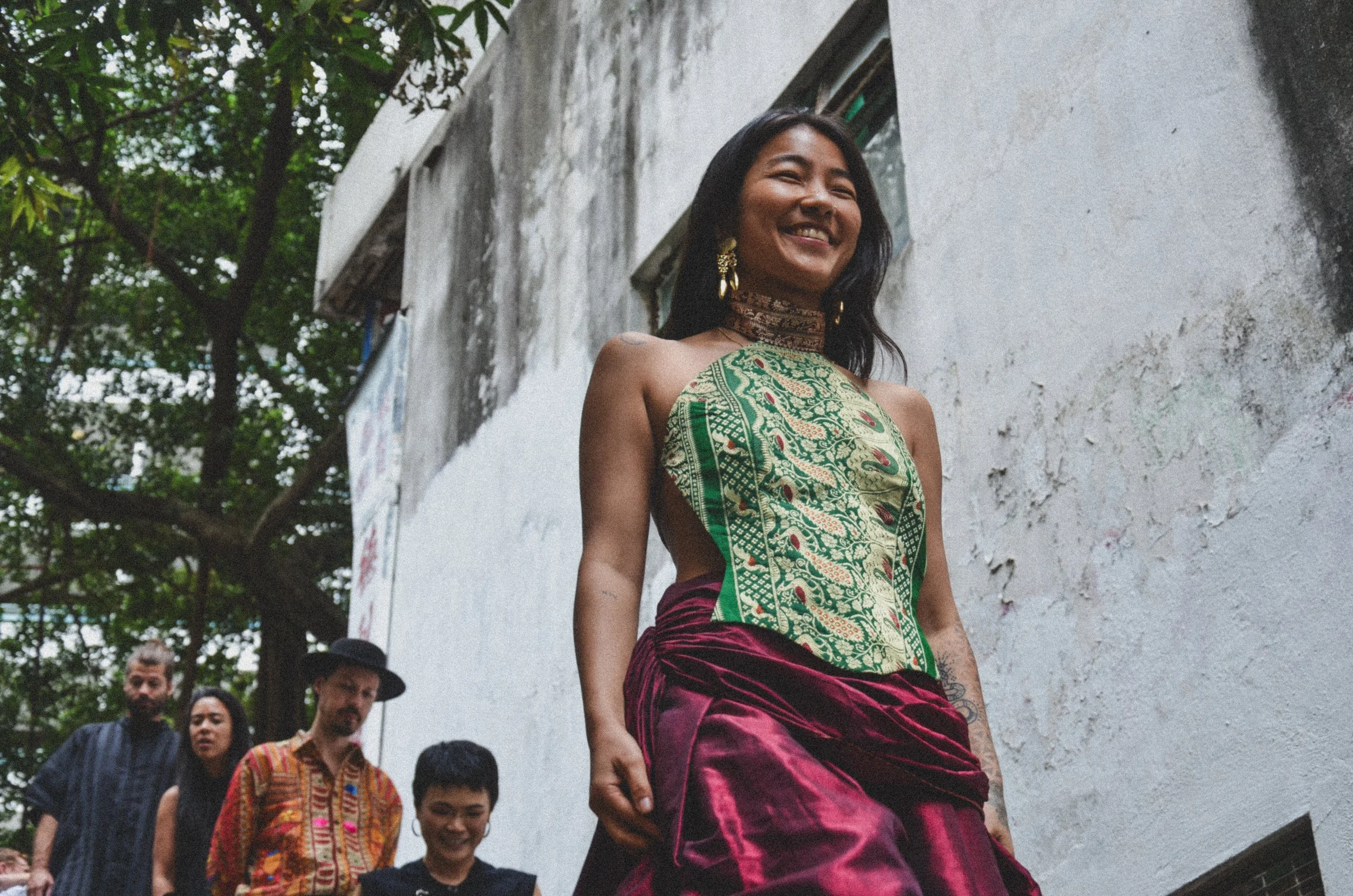 A woman smiling while walking outdoors with a group of people, wearing traditional clothing, against a weathered white wall and surrounded by trees.