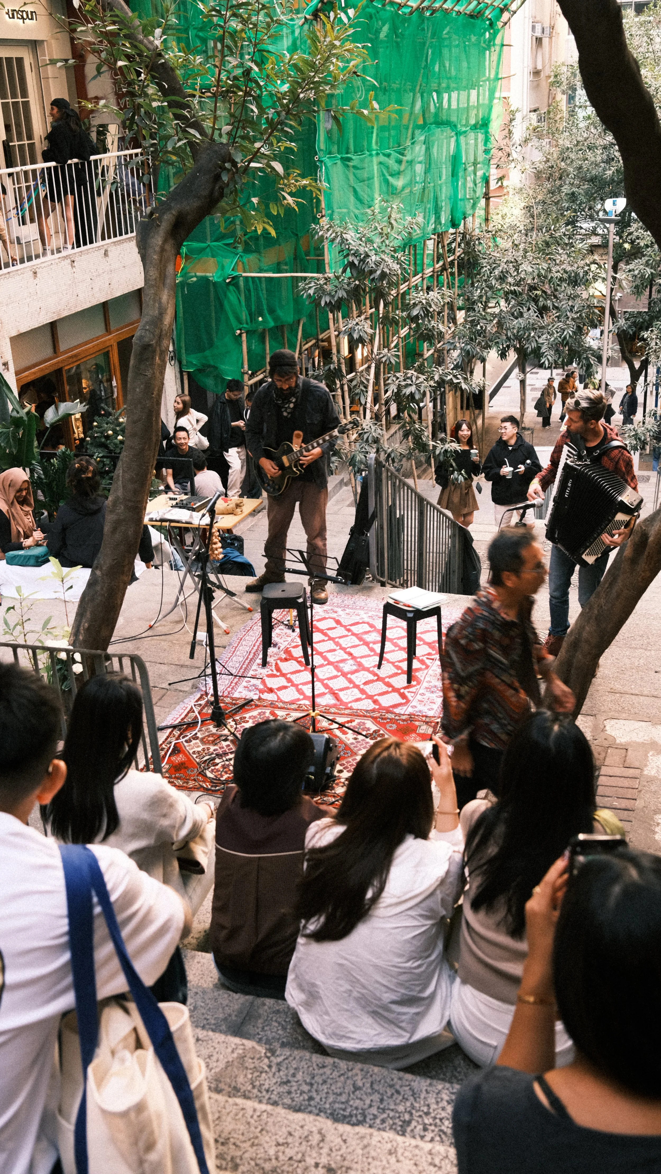 Street performance with musicians playing guitar and accordion on a small stage with an audience seated and standing around in an outdoor urban setting, surrounded by trees and building facades.
