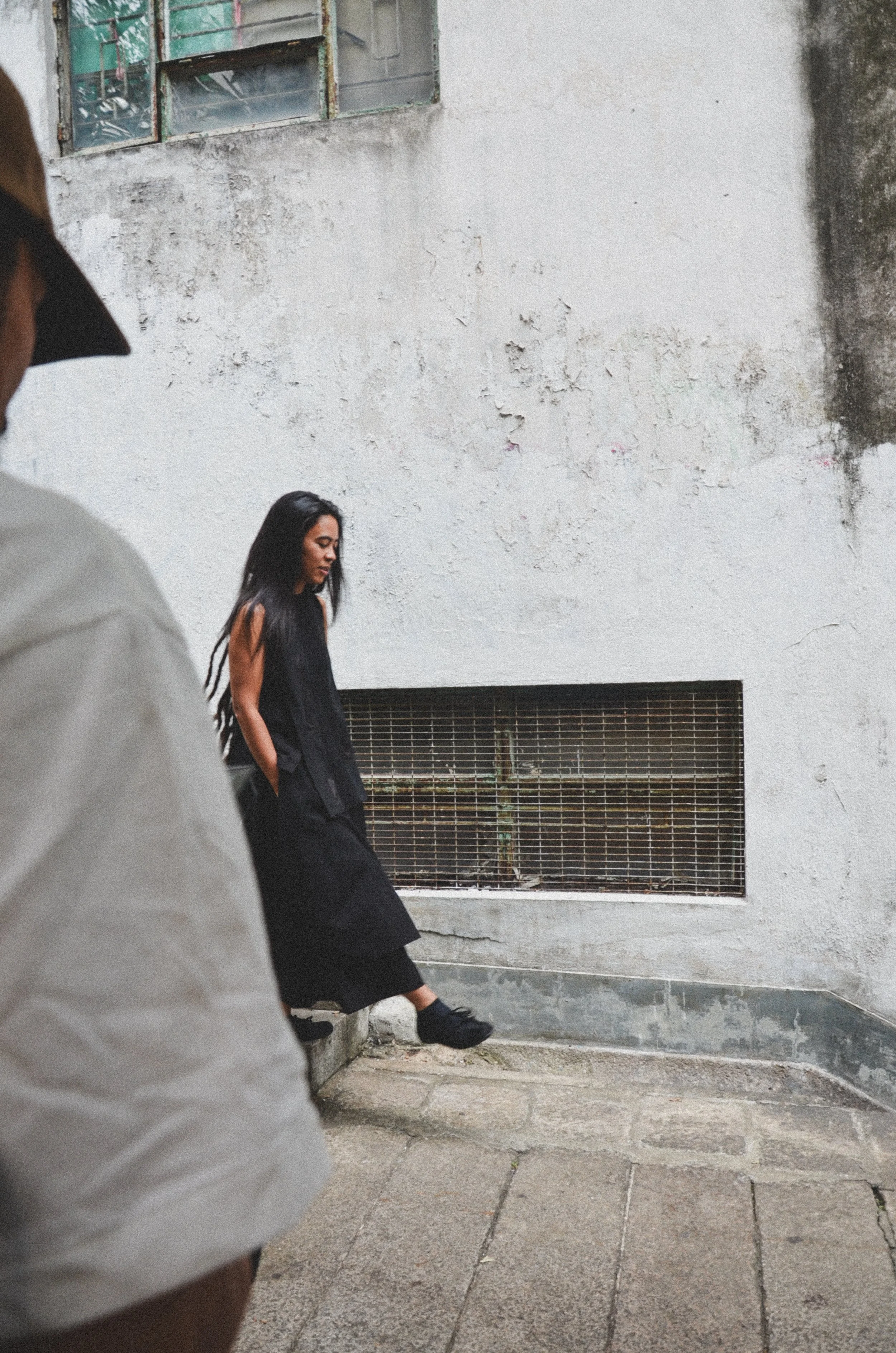 A woman with long dark hair wearing a black dress walking on a concrete sidewalk near a textured white wall with a metal vent, with a person in a gray shirt and wide-brimmed hat partially visible in the foreground.