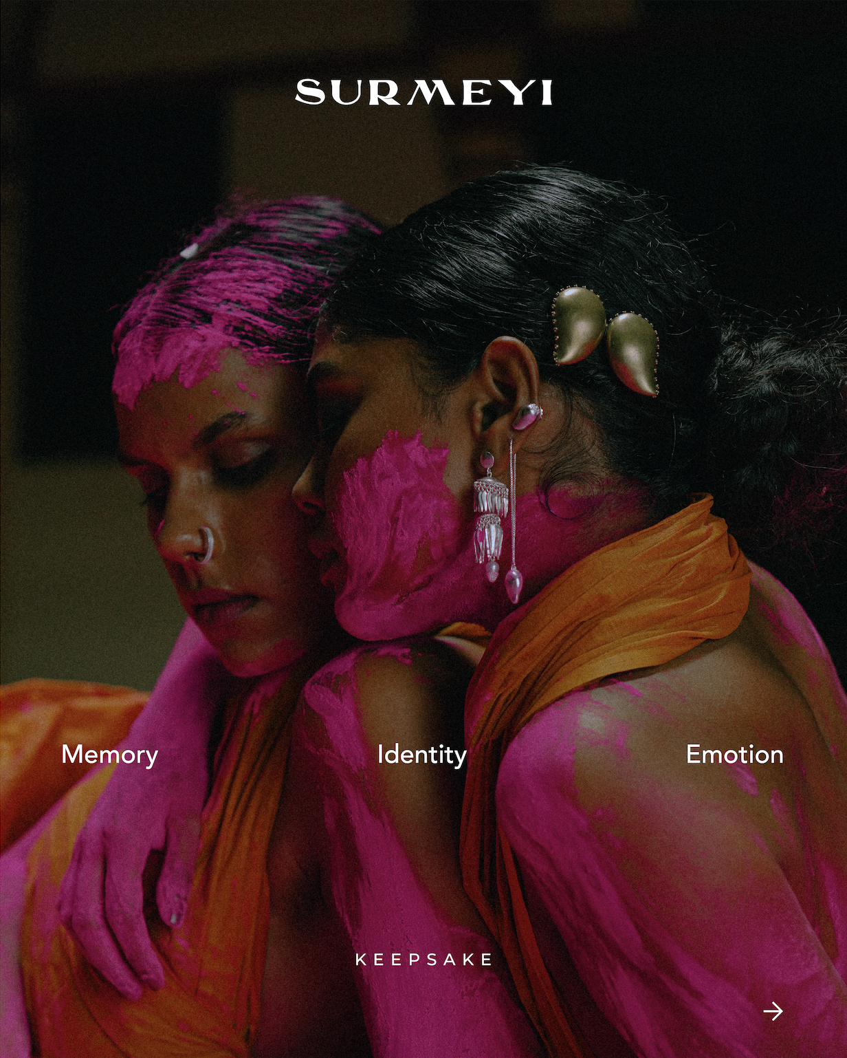 Close-up of two women embracing, with pink paint on their faces and bodies, wearing colorful earrings and headpieces. The image has the text 'SURMEYI' at the top and 'Memory,' 'Identity,' 'Emotion,' and 'KEEPSAKE' at the bottom.