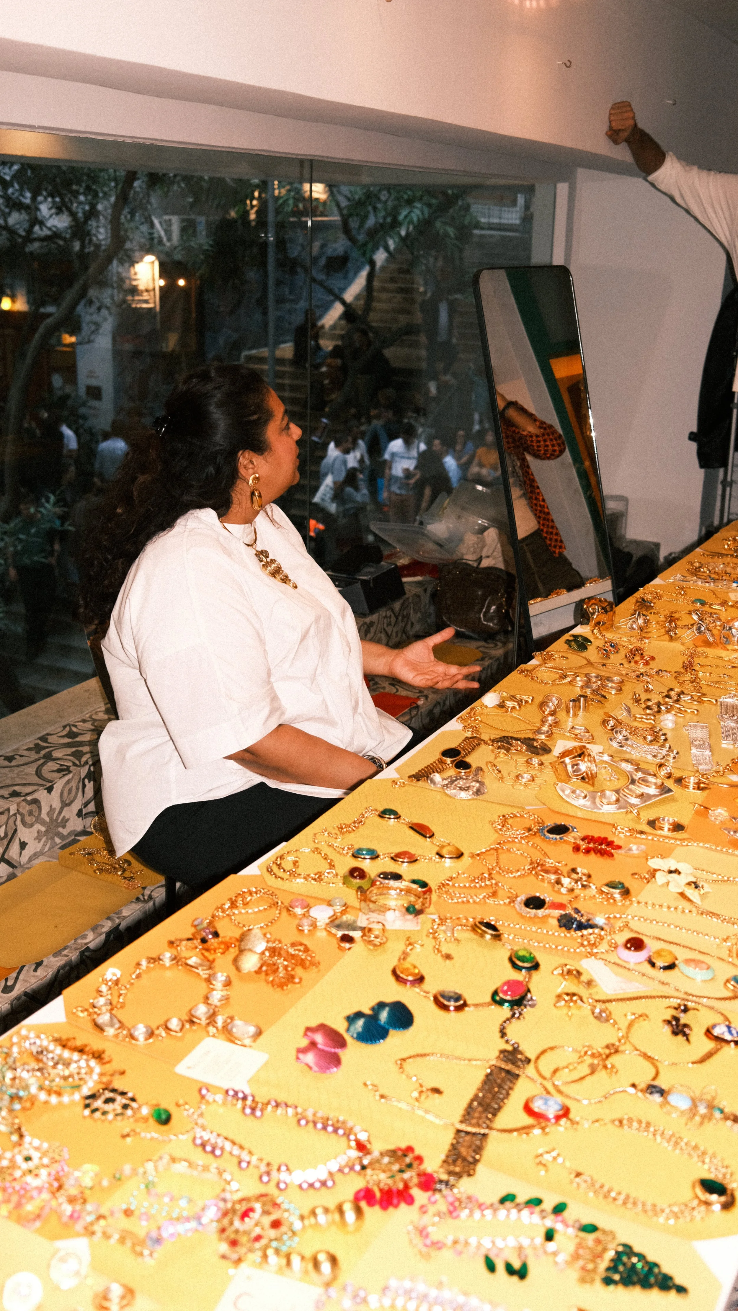 Woman sitting at a table covered with various jewelry, talking to someone in a white top, in a room with large windows showing people outside.