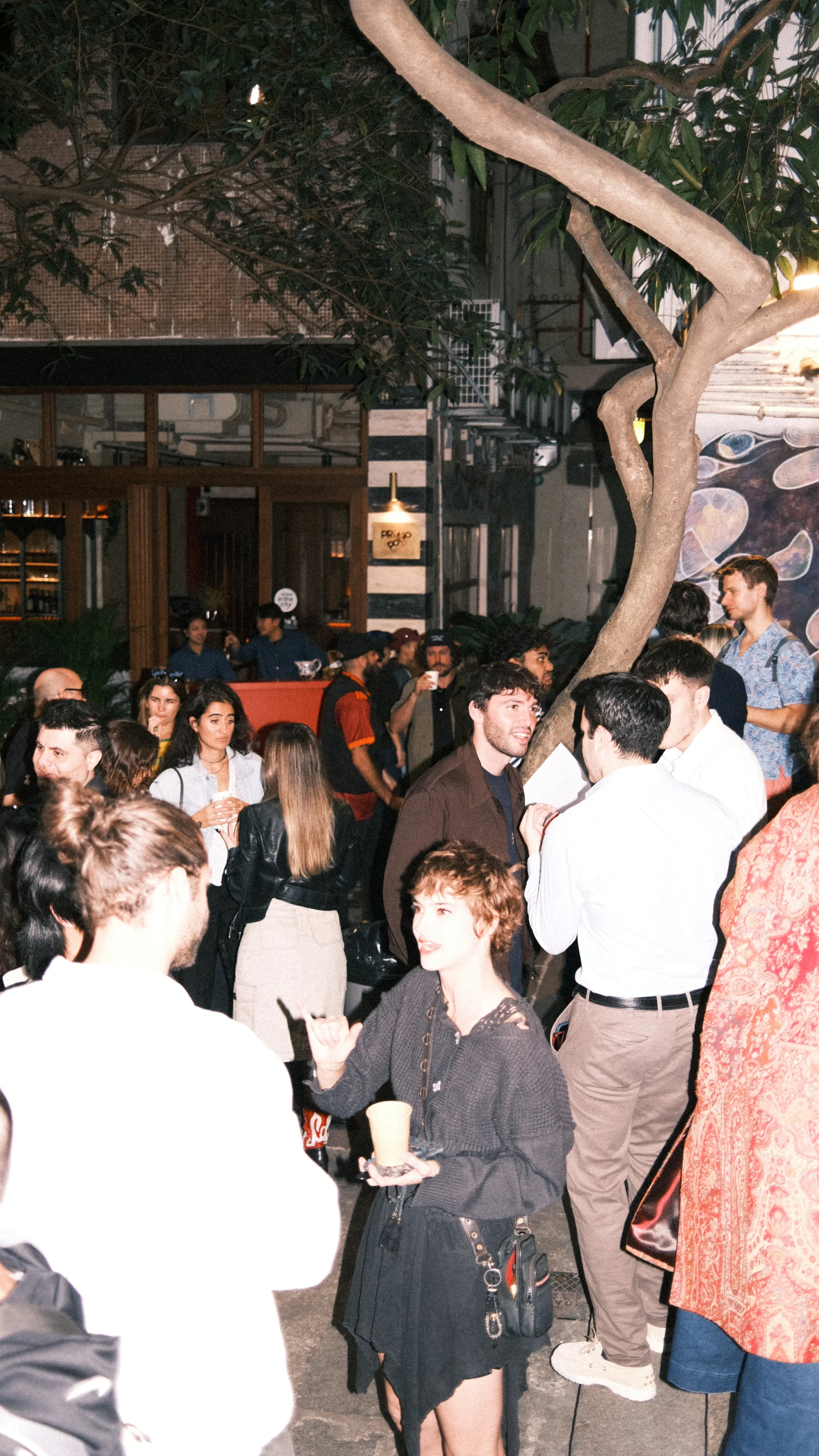 People socializing and mingling at an indoor venue, with a large tree and mural art in the background.
