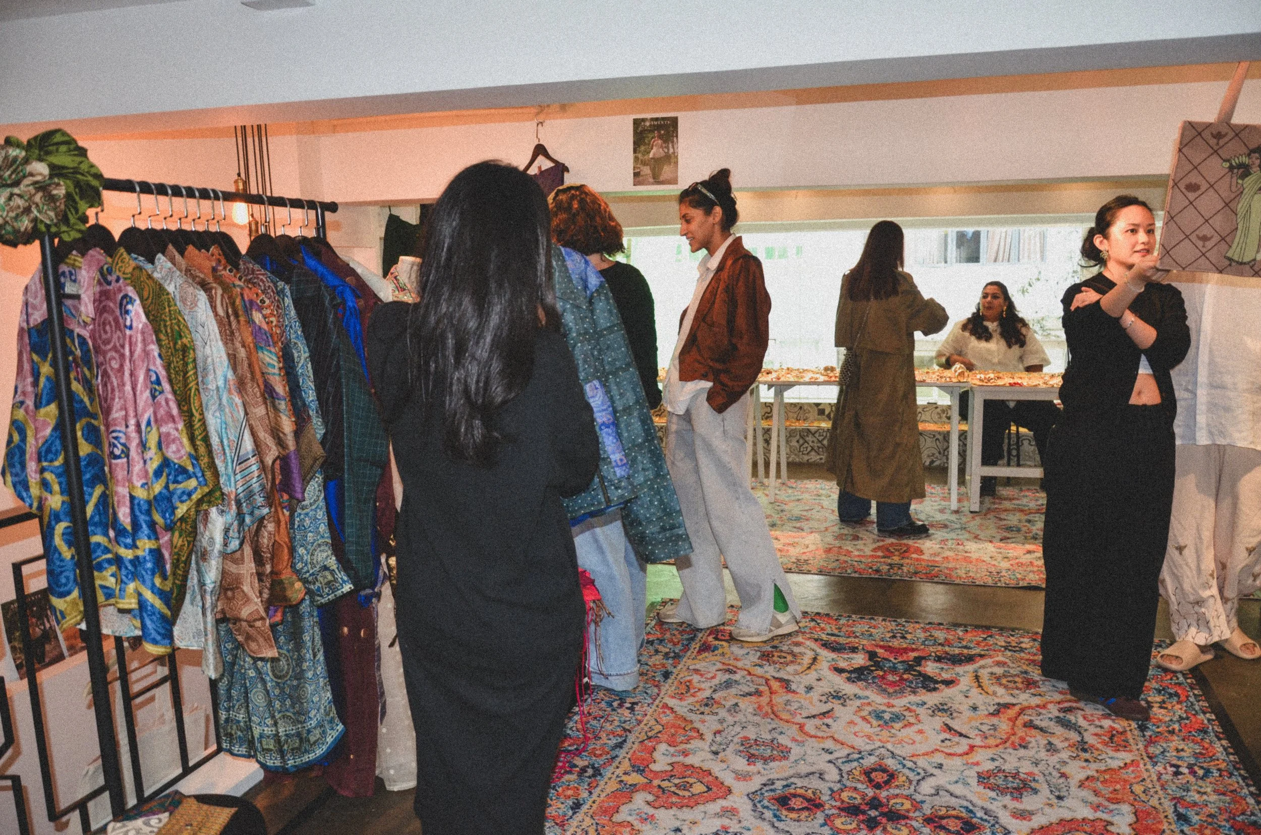 People shopping inside a vintage clothing store with colorful garments on racks, a patterned carpet, and a natural light window.