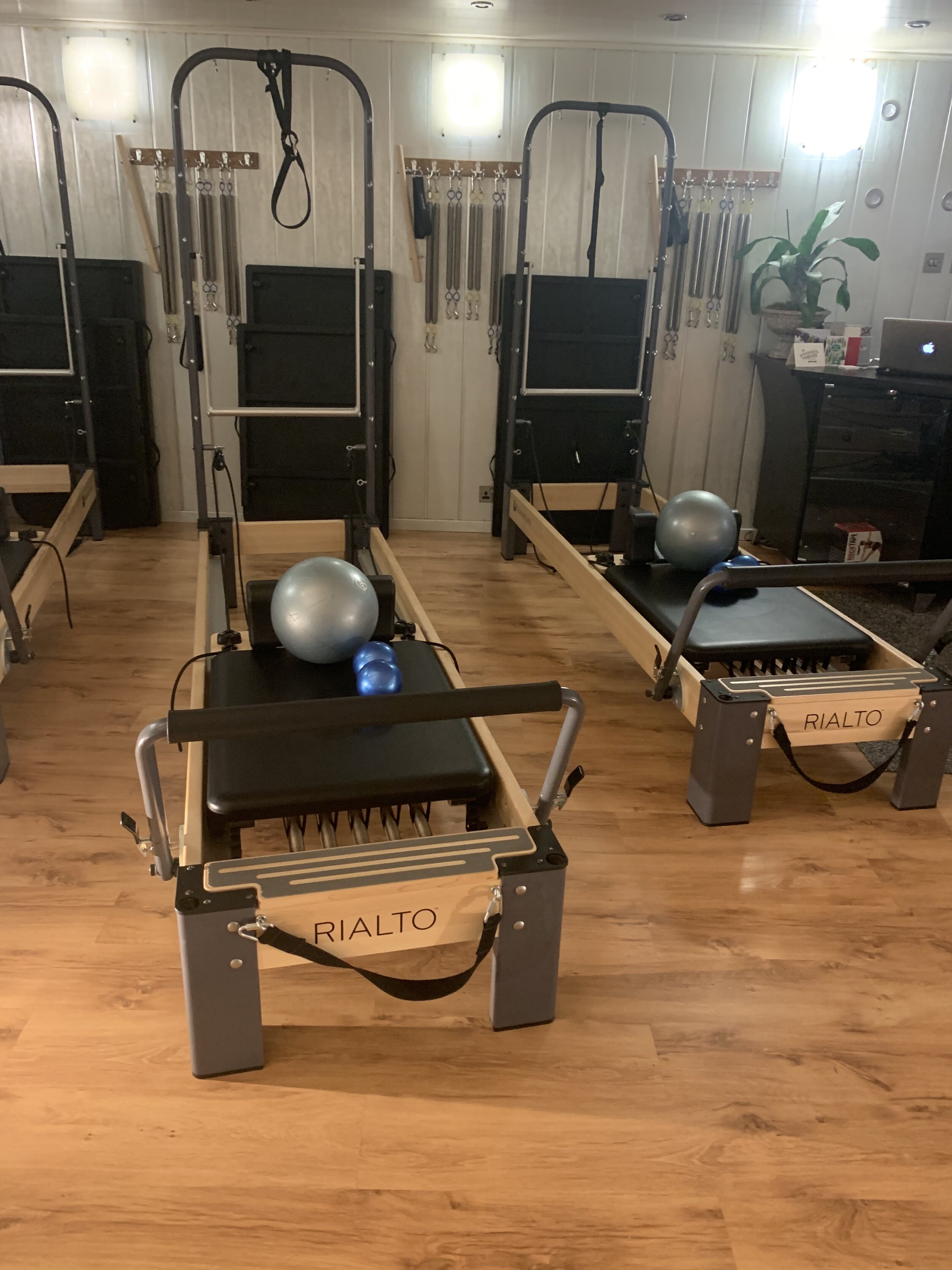 Two Pilates reformer machines with exercise balls, blue and silver, placed on top, in a fitness studio with wooden flooring and wall-mounted resistance bands in the background.