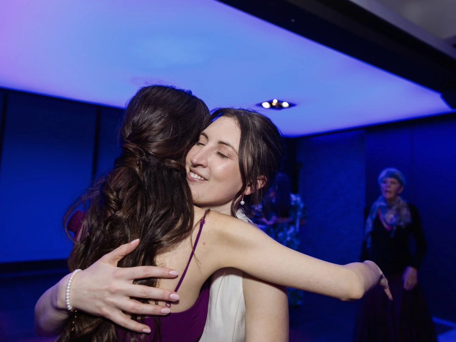 Bride hugging friend with her eyes closed against neon blue light.